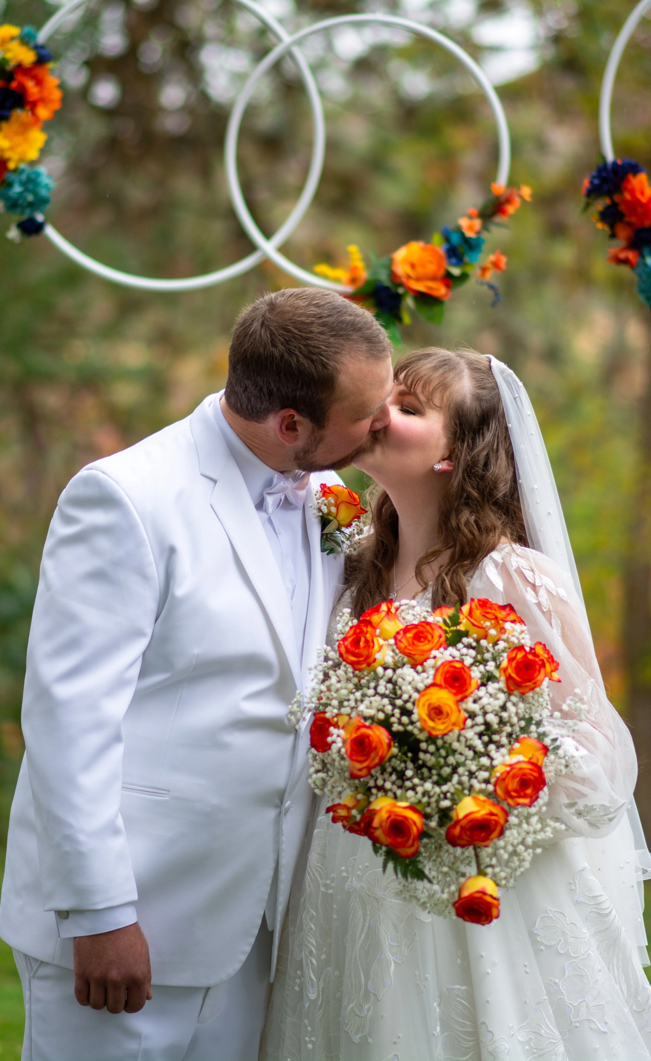 A couple in wedding attire sharing a kiss outdoors, with colorful circular floral decorations hanging above them.