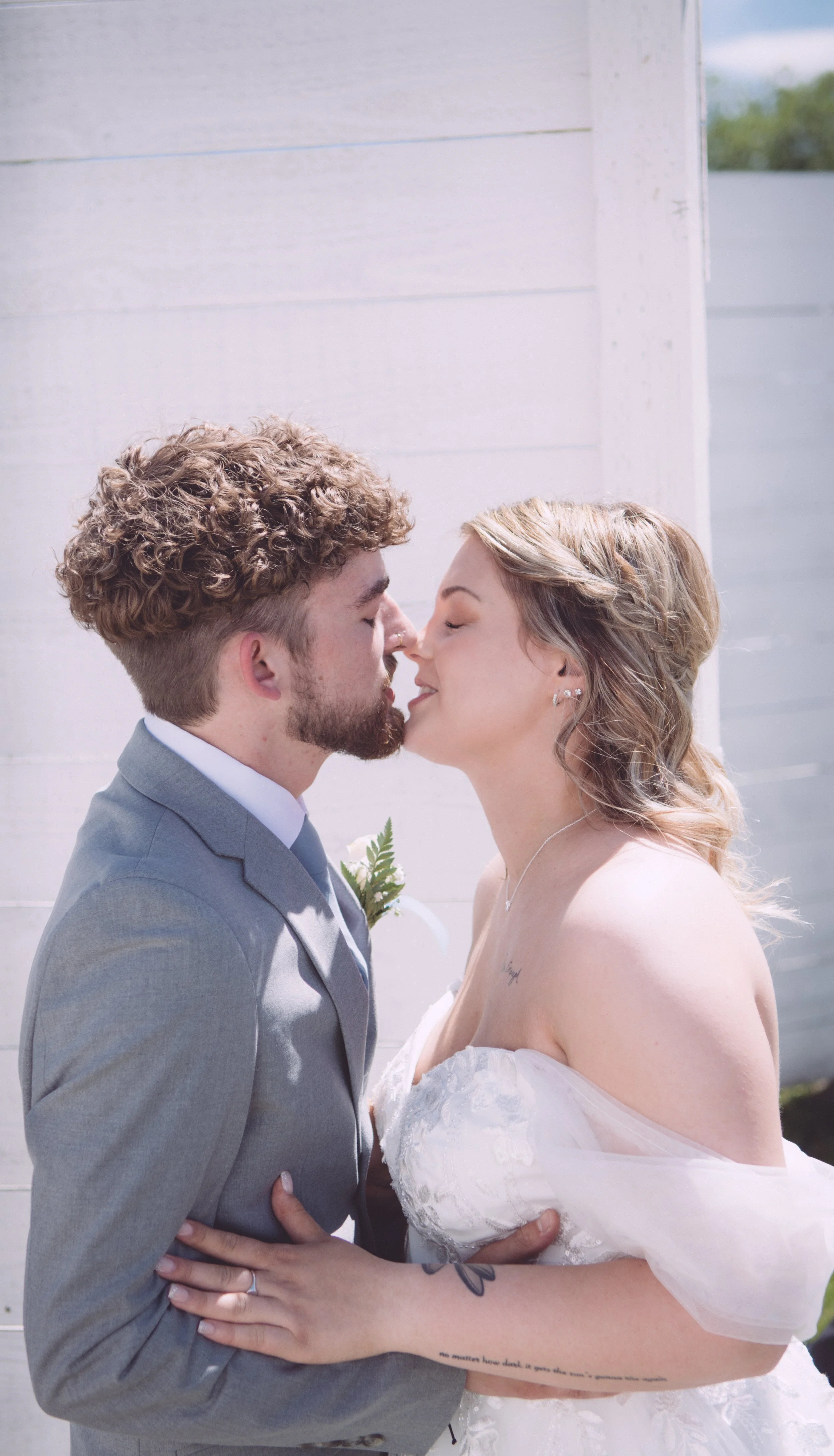 A newlywed couple with eyes closed and noses touching, in wedding attire outdoors in front of a white wall.
