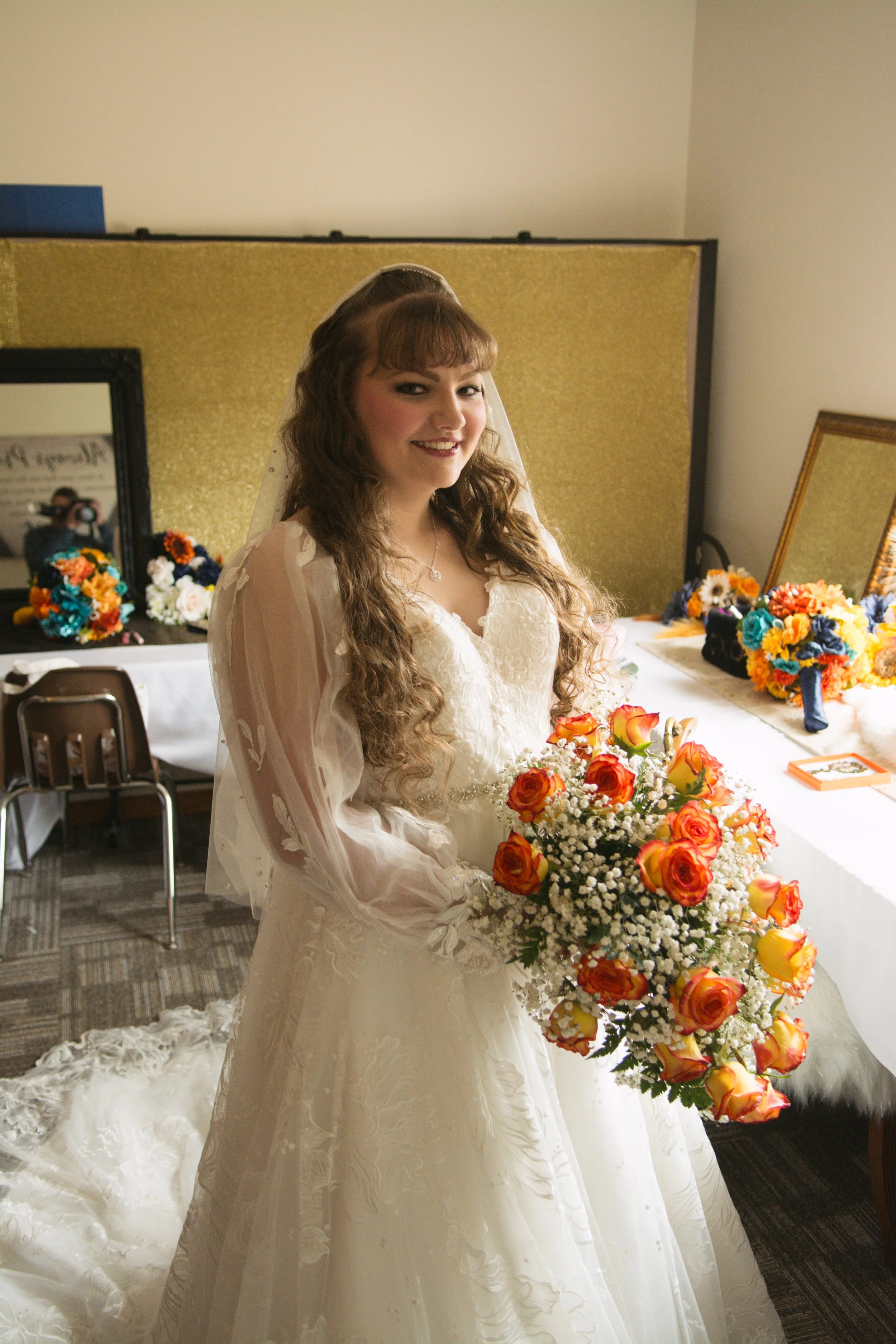 A bride in a white wedding dress holding a bouquet of orange roses and white baby's breath, standing indoors with a table of floral arrangements and a mirror behind her.