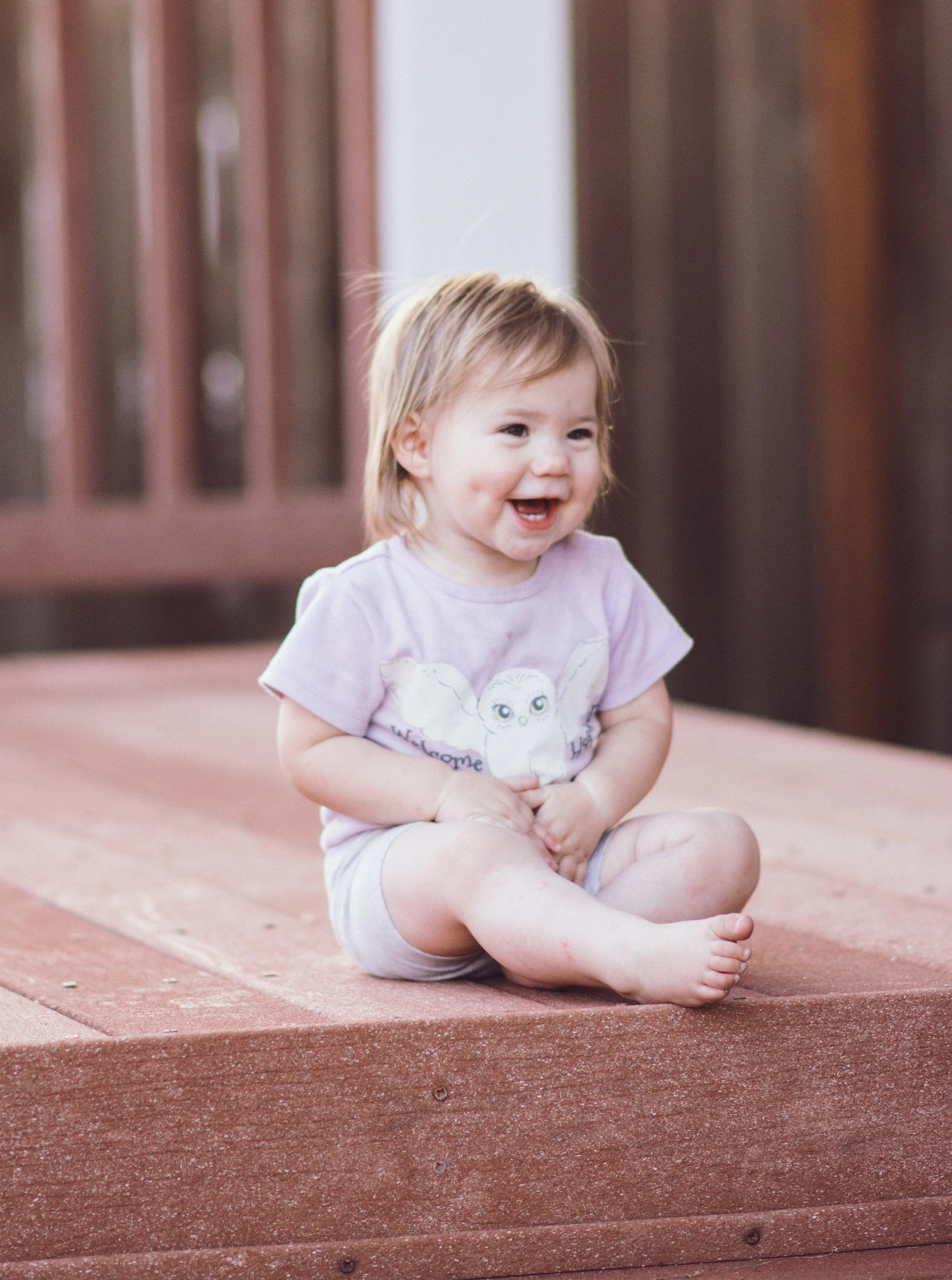 A laughing toddler girl with light brown hair sitting on a wooden deck, wearing a light purple shirt and light shorts.