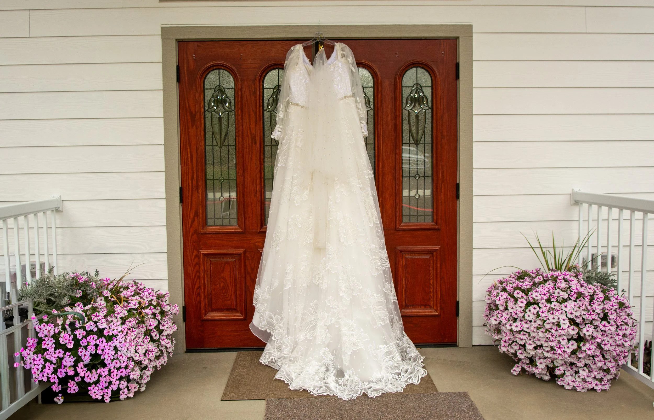 A wedding dress hanging on a hanger in front of a wooden front door, with pink and purple petunias on both sides of the entrance.