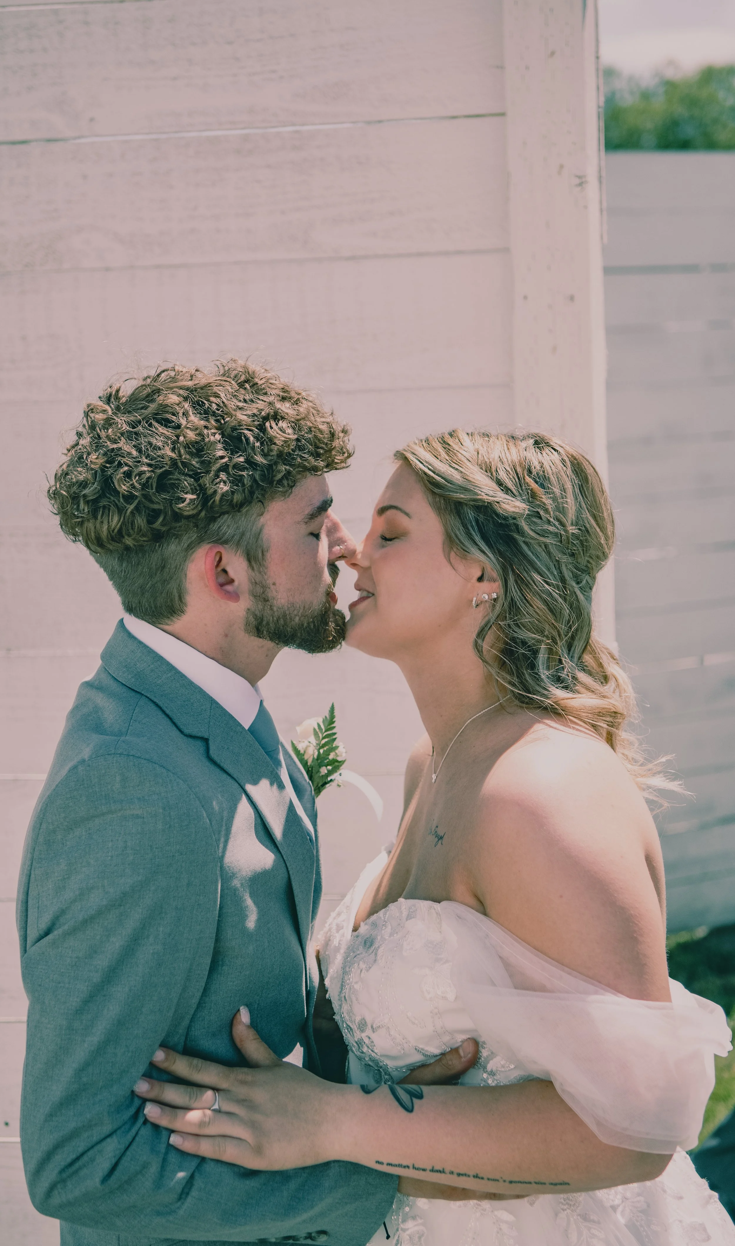 A couple in wedding attire sharing a kiss outdoors against a wooden fence.