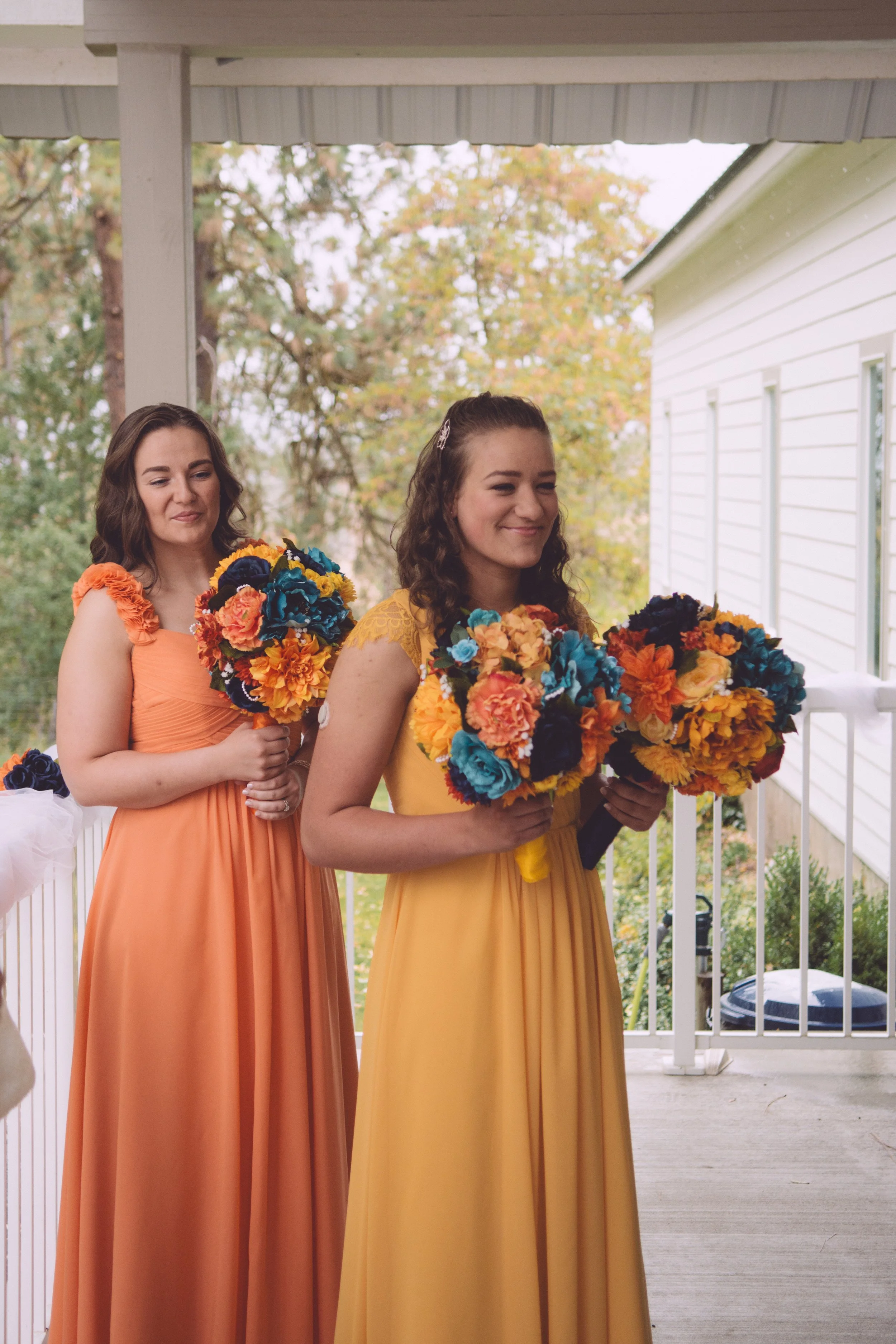 Two women in colorful dresses holding bright flower bouquets, standing on a porch with trees and a white house in the background.