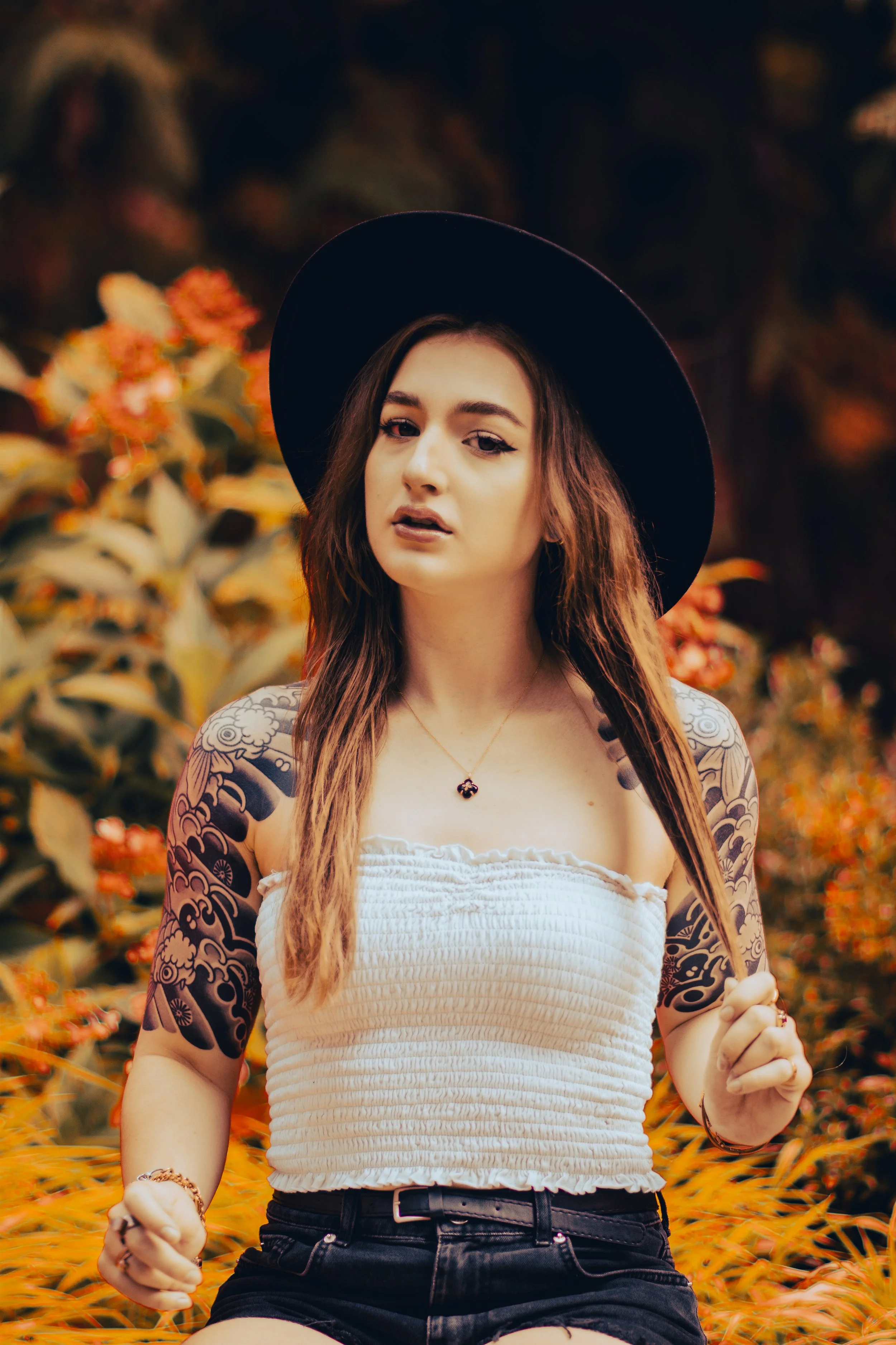 A young woman with long brown hair, wearing a black wide-brimmed hat, a white strapless top, and black shorts, standing outdoors among orange and yellow foliage, with visible tattoos on her arms.
