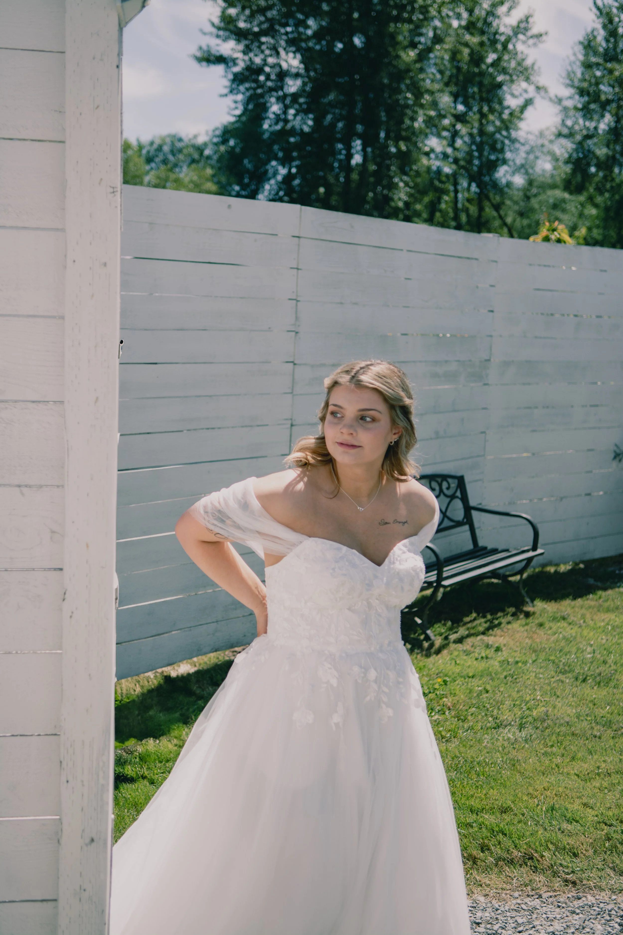 A bride in a white wedding gown with floral embroidery, standing outdoors near a white wooden fence and a park bench, with greenery and trees in the background.