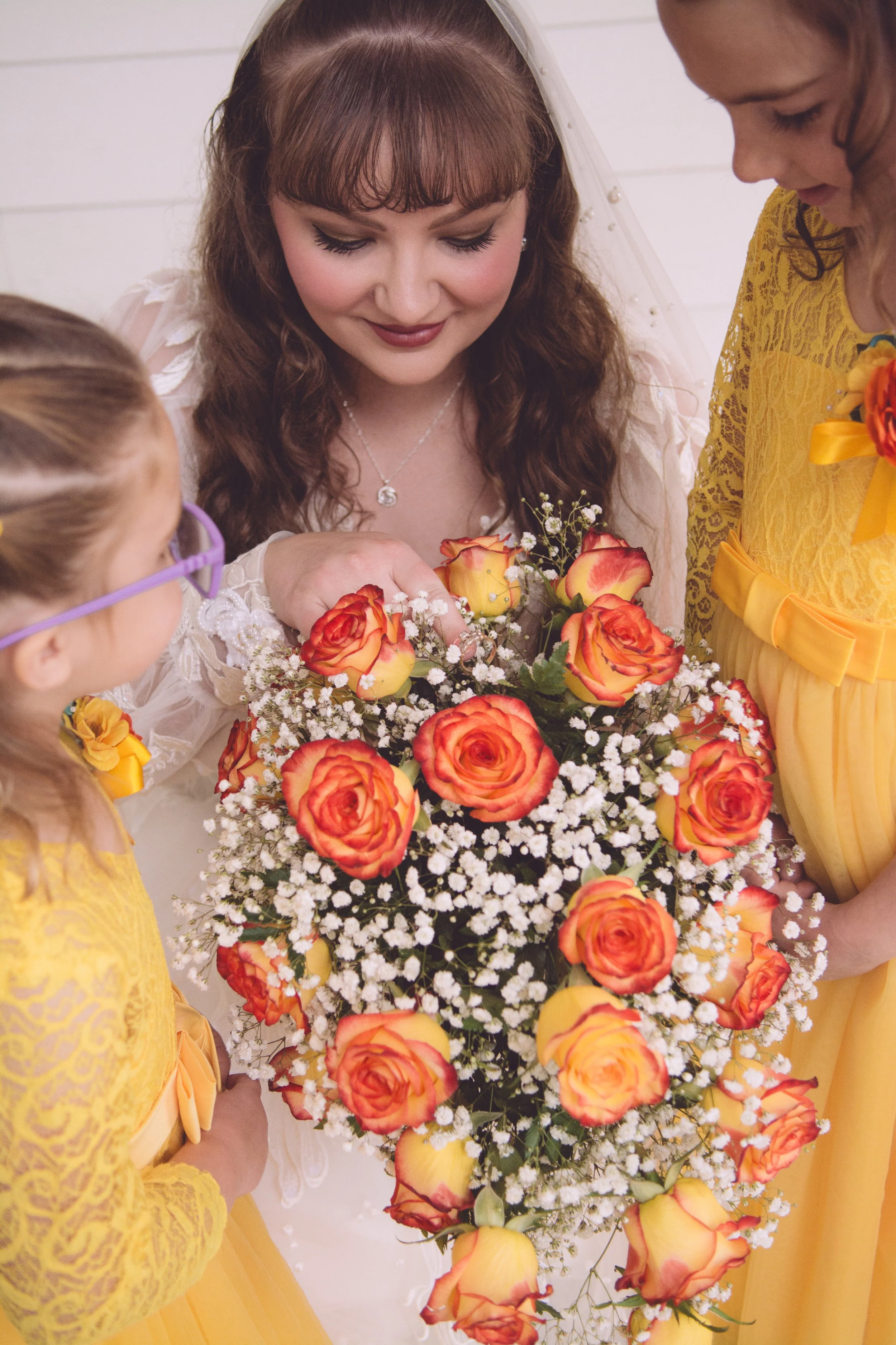 A bride holding a bouquet of orange roses and white baby's breath, surrounded by two young girls in yellow dresses during a wedding ceremony.