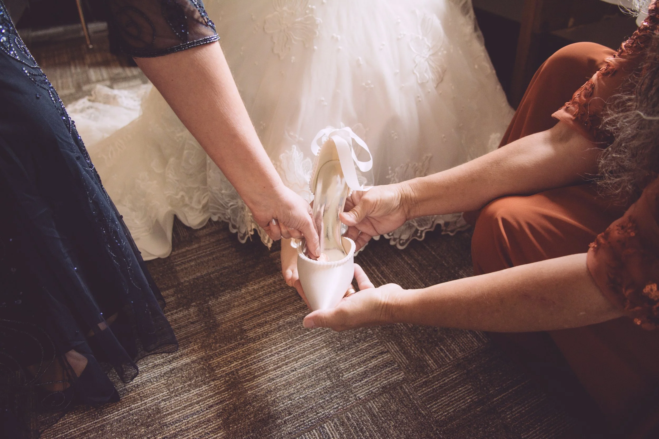 Two women are holding a white ballet shoe with a ribbon in between, with a woman in a wedding dress in the background.