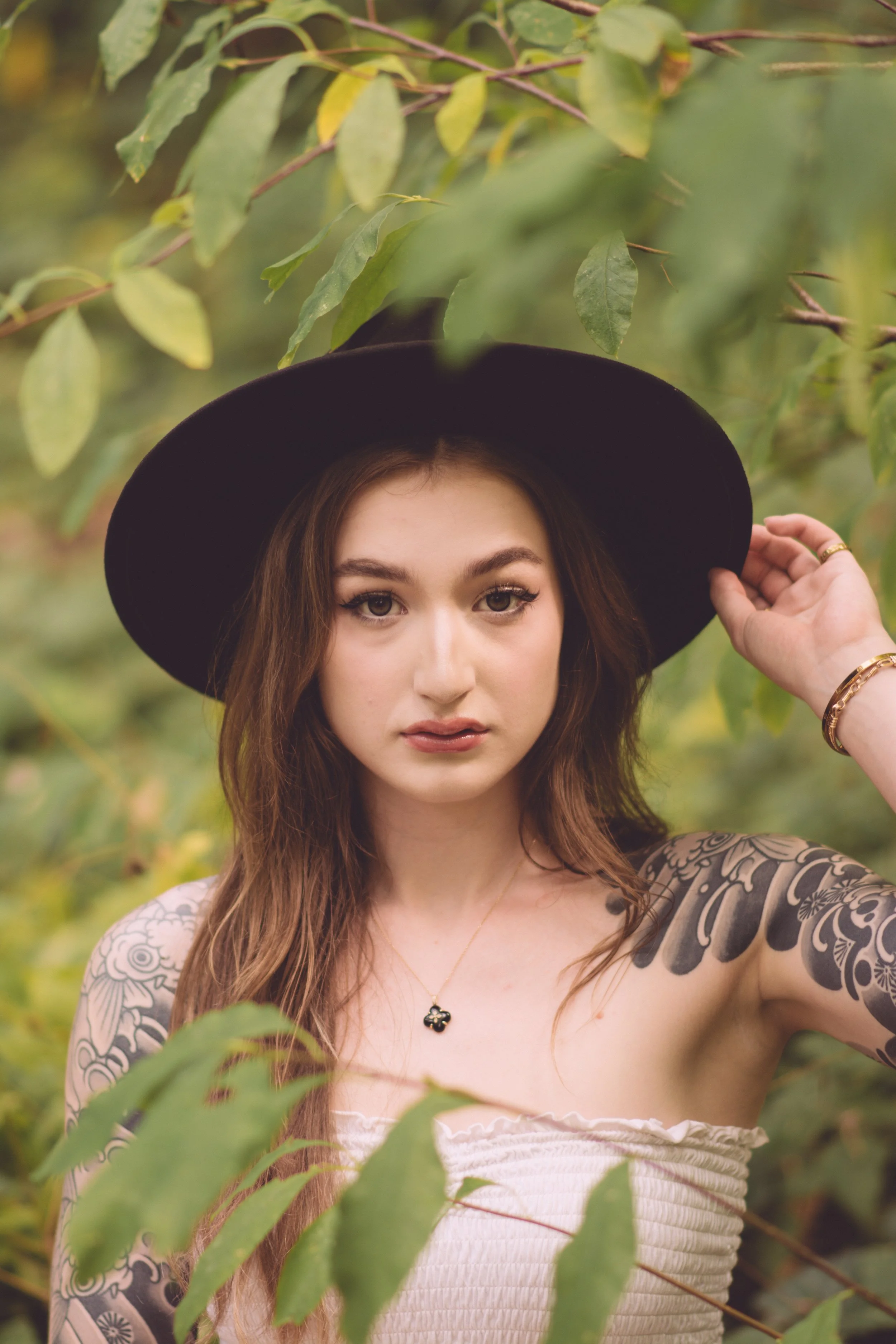 Young woman with long brown hair wearing a black wide-brim hat, beige strapless top, and jewelry, standing outdoors surrounded by green leaves.