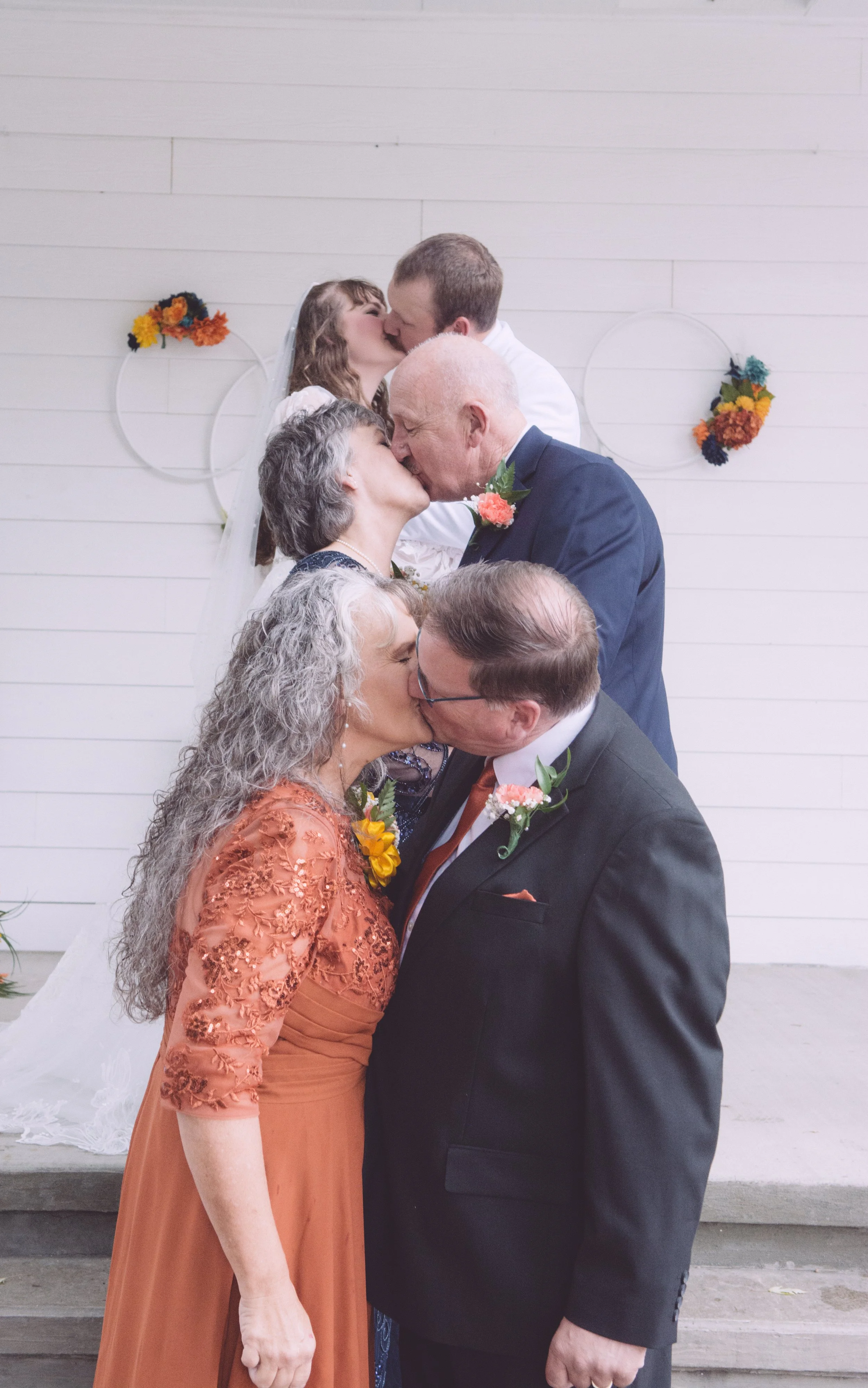 Couples kissing during wedding ceremony, with three couples in the foreground and two couples kissing in the background, against a white wall with circular floral decorations.