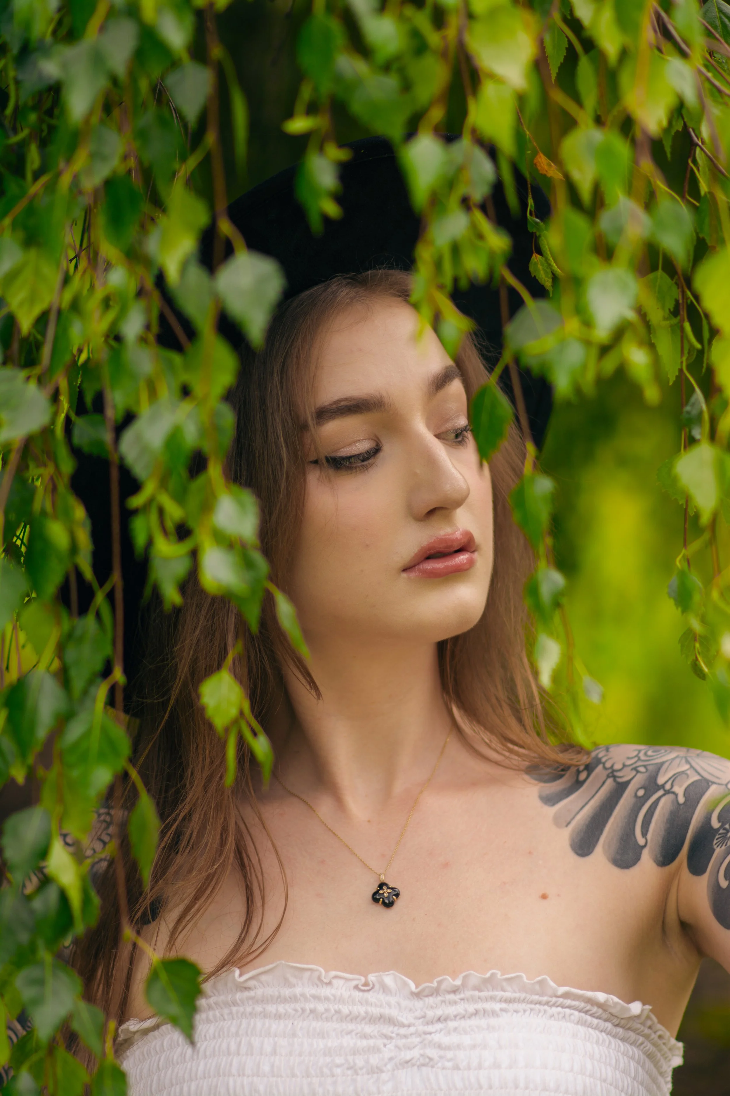A woman with long brown hair and a tattoo on her arm, wearing a black hat and a white strapless top, stands amidst green leaves.