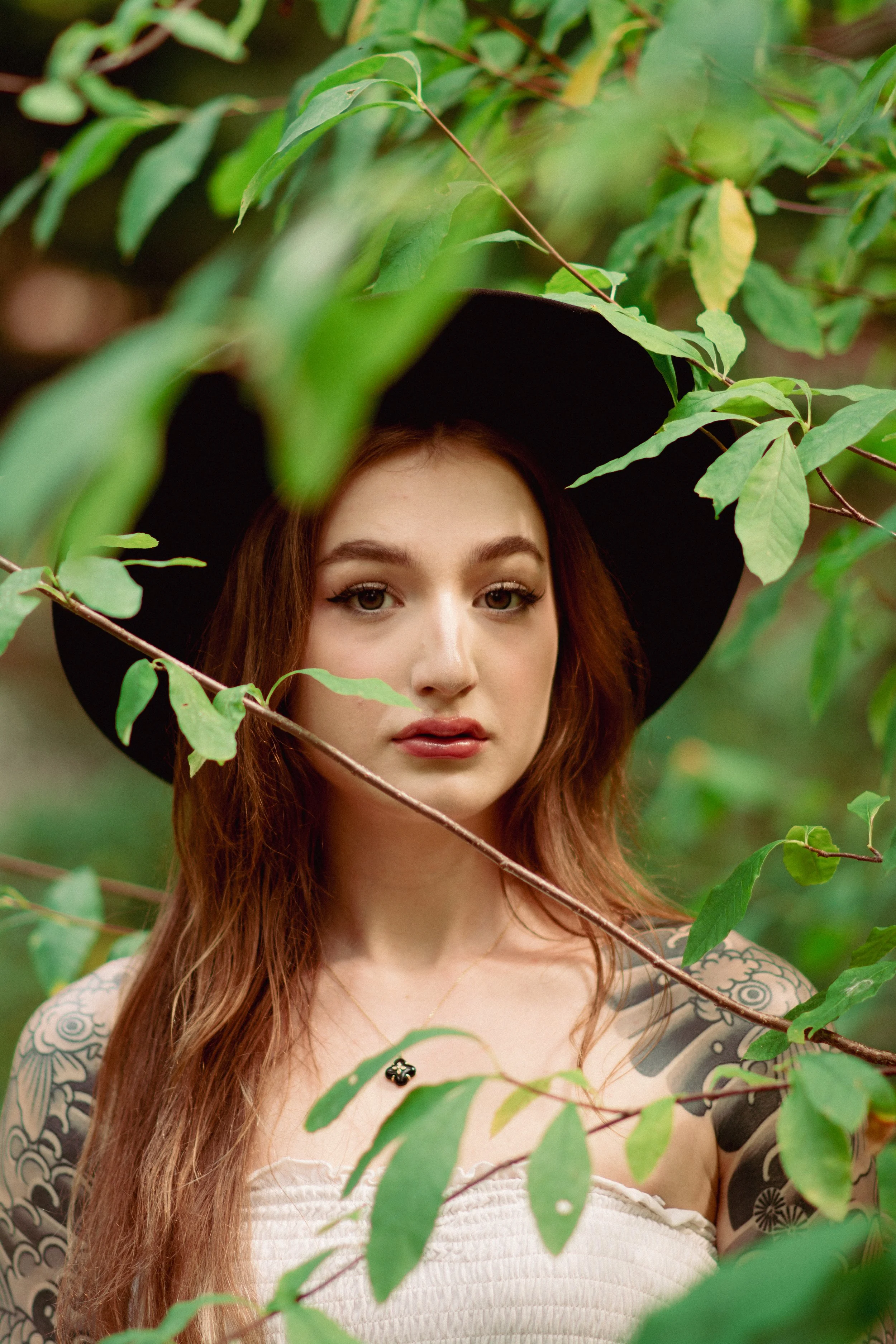 Portrait of a young woman with red hair wearing a black hat and a white top, surrounded by green leaves.