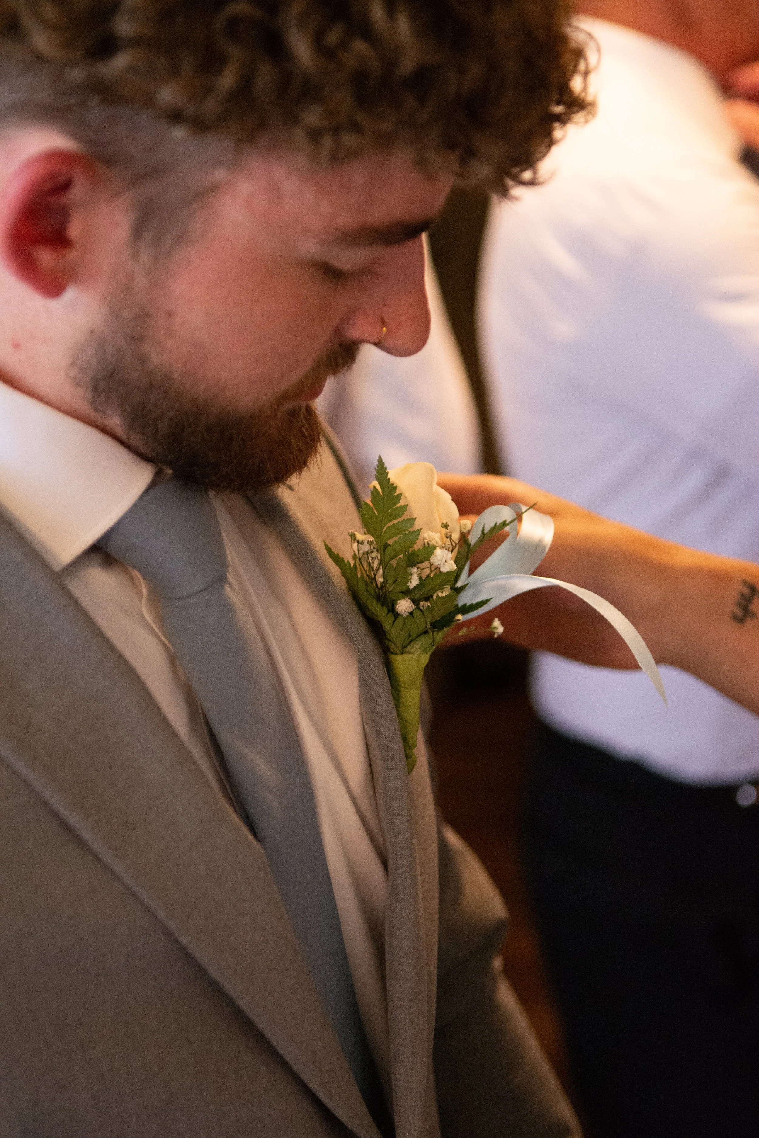 A man with a beard and curly hair, dressed in a beige suit and white shirt, during a wedding ceremony. He has a boutonniere with white flowers, greenery, and a white ribbon pinned to his lapel.