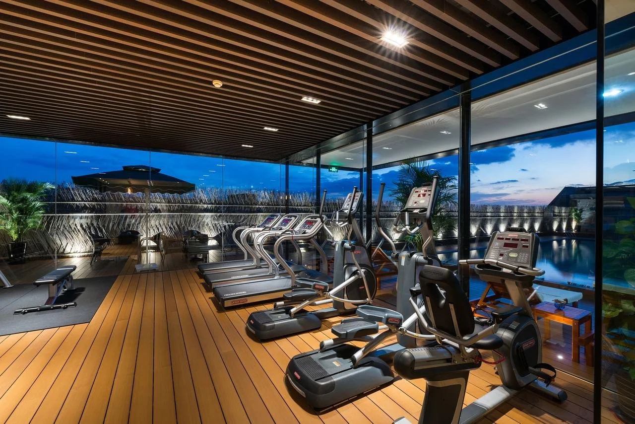 Indoor gym with cardio equipment like treadmills, ellipticals, and stationary bikes, overlooking an outdoor pool and city skyline during dusk.