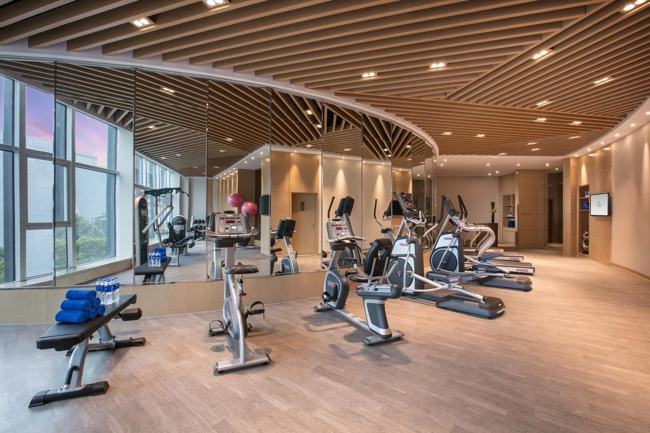 A modern gym with various exercise equipment including stationary bikes, treadmills, and weights. The gym has large windows, wooden ceiling panels, and mirrors on the wall.