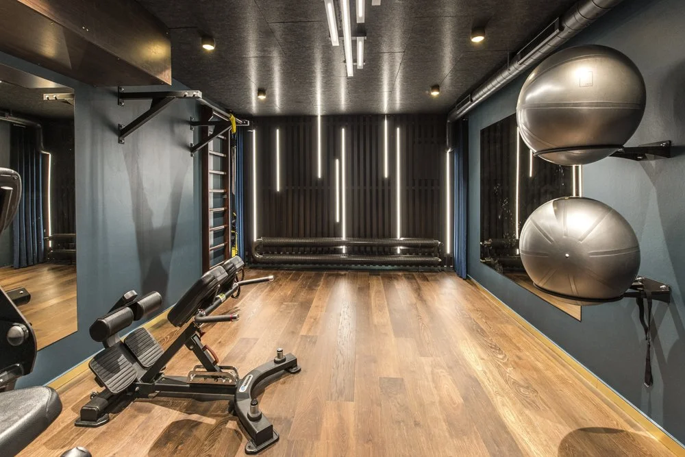 Empty home gym with wooden flooring, black wall with vertical light strips, exercise equipment including balance balls on the wall, a mirror, and a wall-mounted pull-up bar.