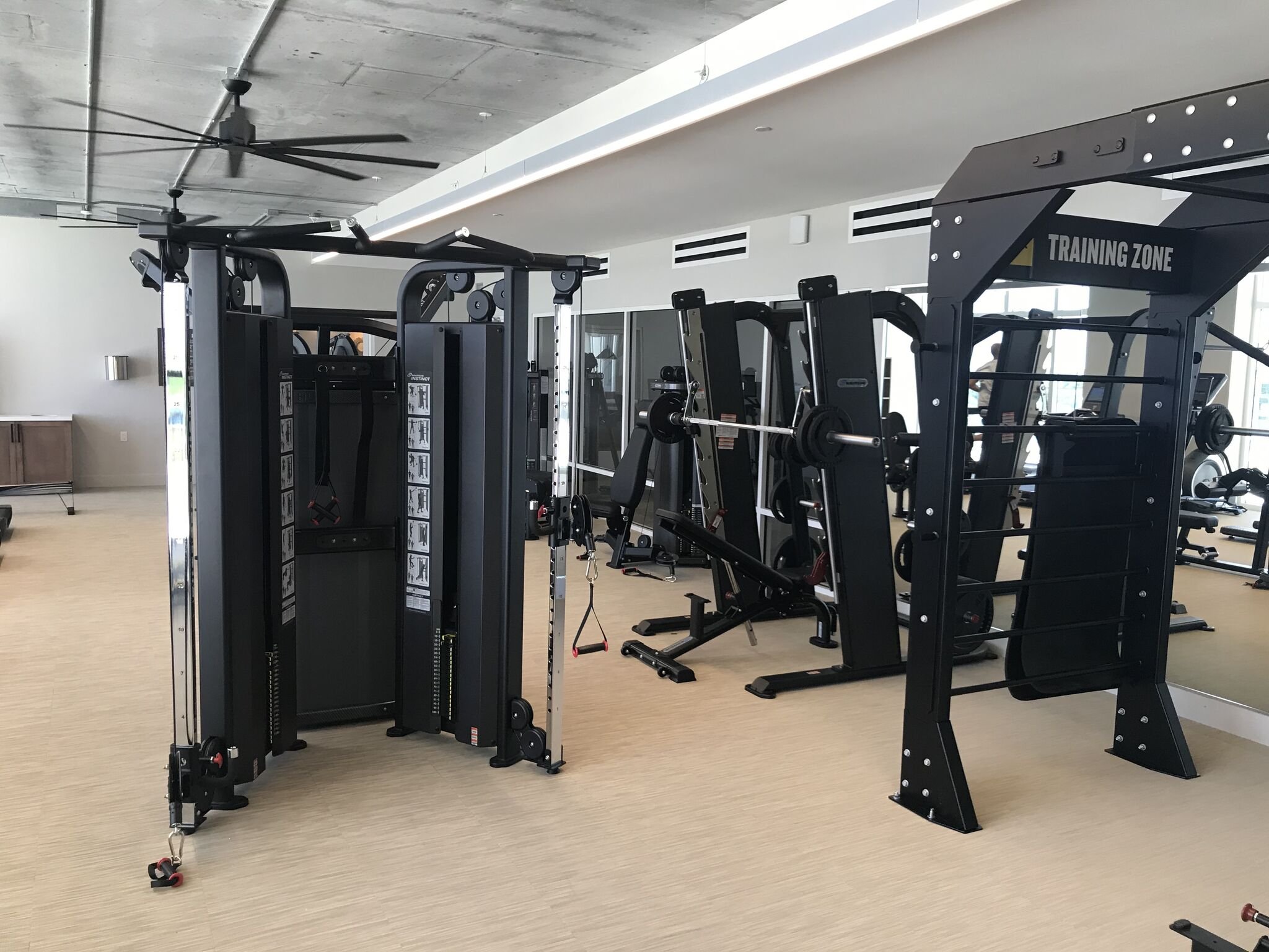 A modern gym with various weightlifting and exercise machines, open space, light wood flooring, large windows, and a ceiling fan.