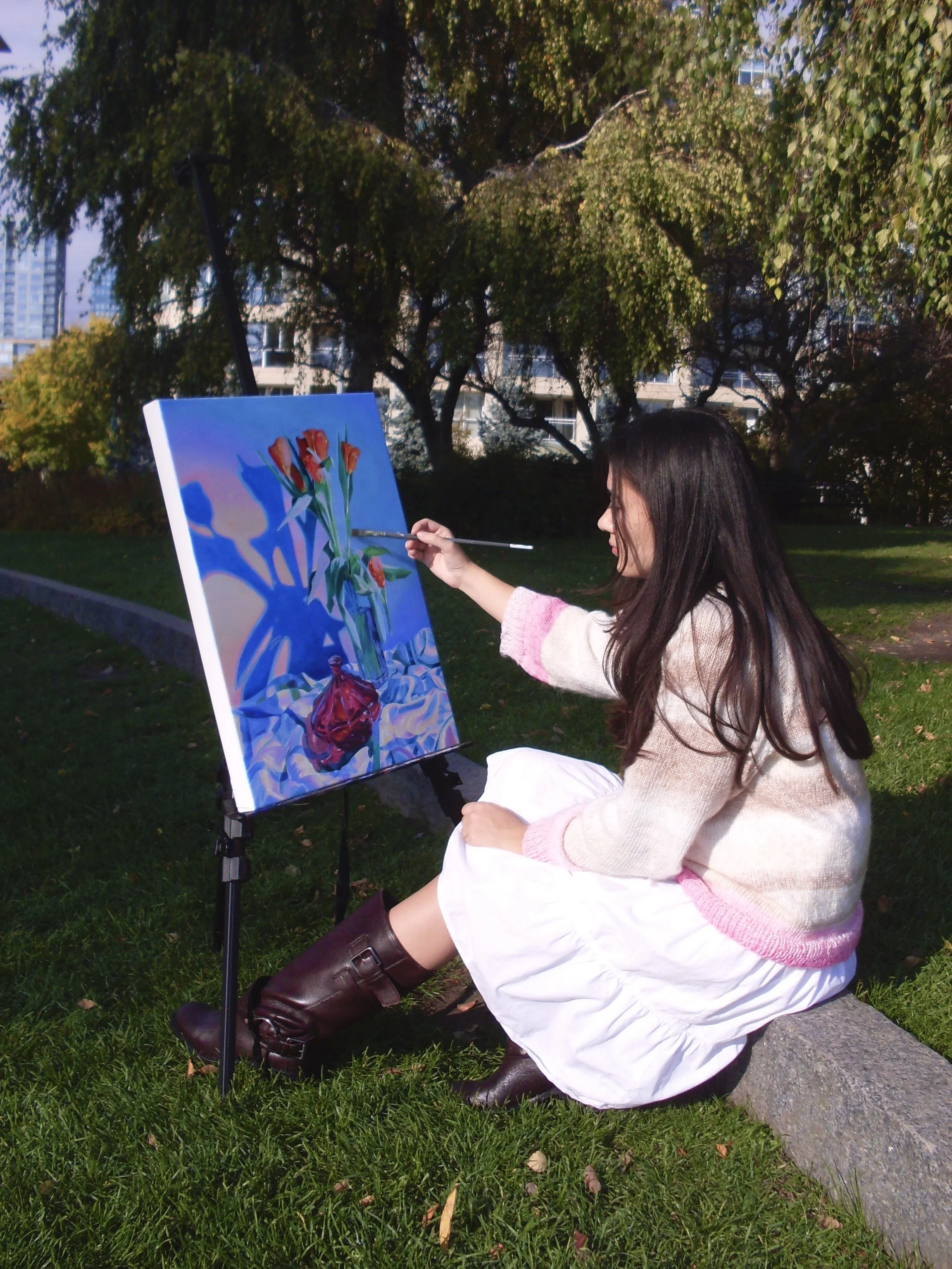 A woman sitting on a stone in a park, painting a still life of flowers in a vase on an easel. She has long dark hair and is wearing a pink and white sweater, a white skirt, and brown boots. Trees and buildings are visible in the background.