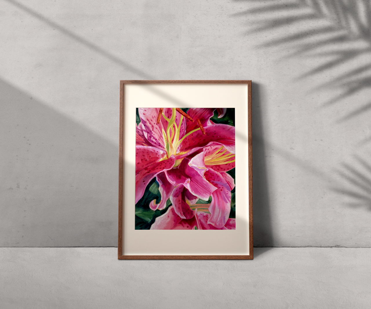 Framed pink lily flower artwork leaning against a light grey wall with shadow of a plant cast on the wall.