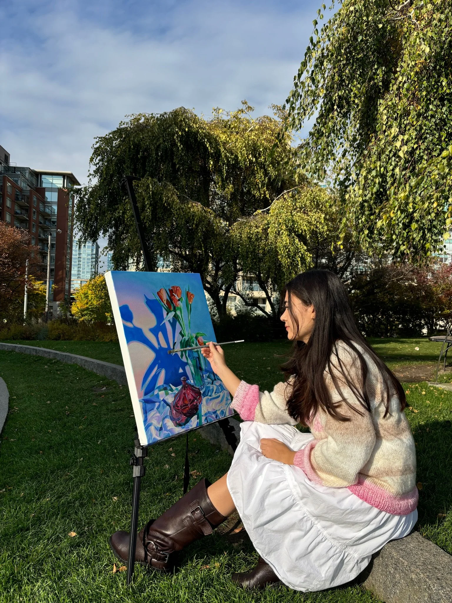 A woman painting a still life of flowers on a canvas outdoors in a park with green grass, trees, and city buildings in the background on a partly cloudy day.