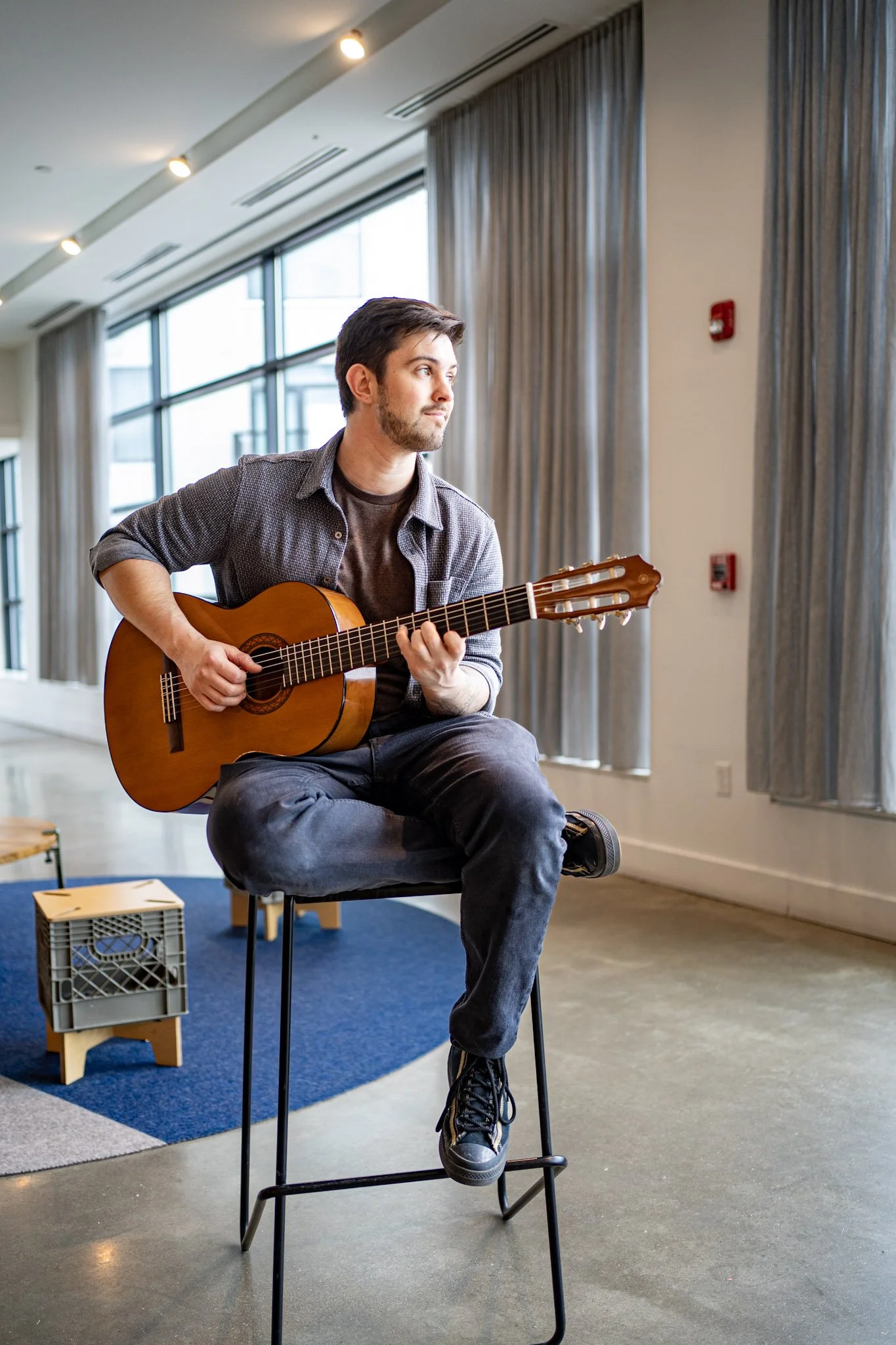 A man playing an acoustic guitar while sitting on a stool in a well-lit room with large windows and curtains.