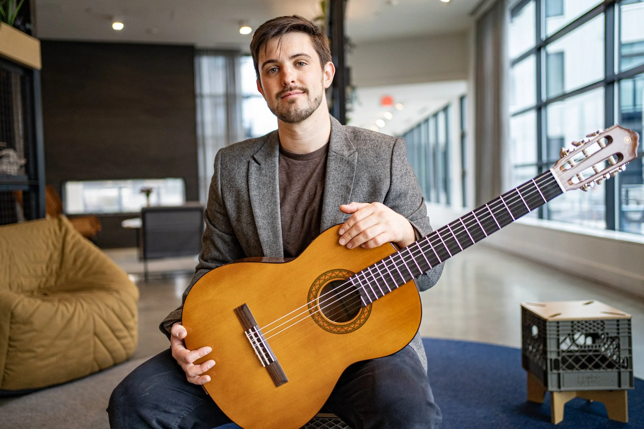 A man with brown hair and a beard holding an acoustic guitar inside a modern, well-lit room with large windows.