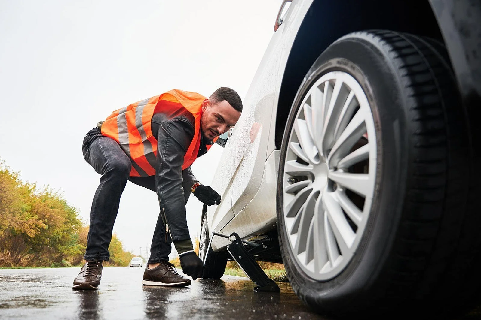 Man in a safety vest changing a flat tire on a car on a wet road with trees in the background.