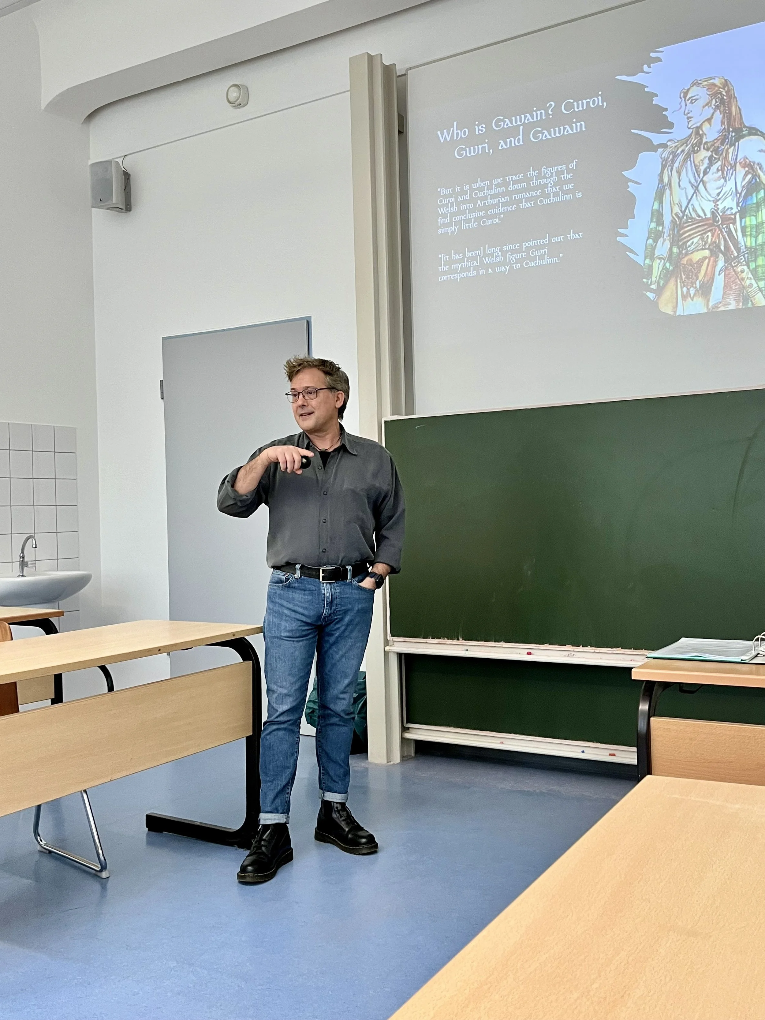 A university classroom with a male speaker standing in front of a blackboard and a projector screen displaying a presentation about Gawain, a character from Welsh mythology. The classroom has wooden desks, a sink, and a white door.