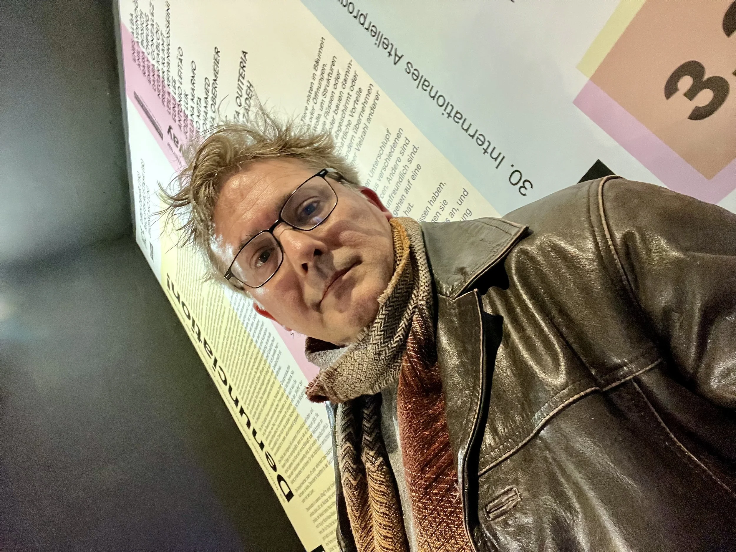 A man with glasses and tousled brown and grey hair taking a selfie in front of a colorful informational poster.