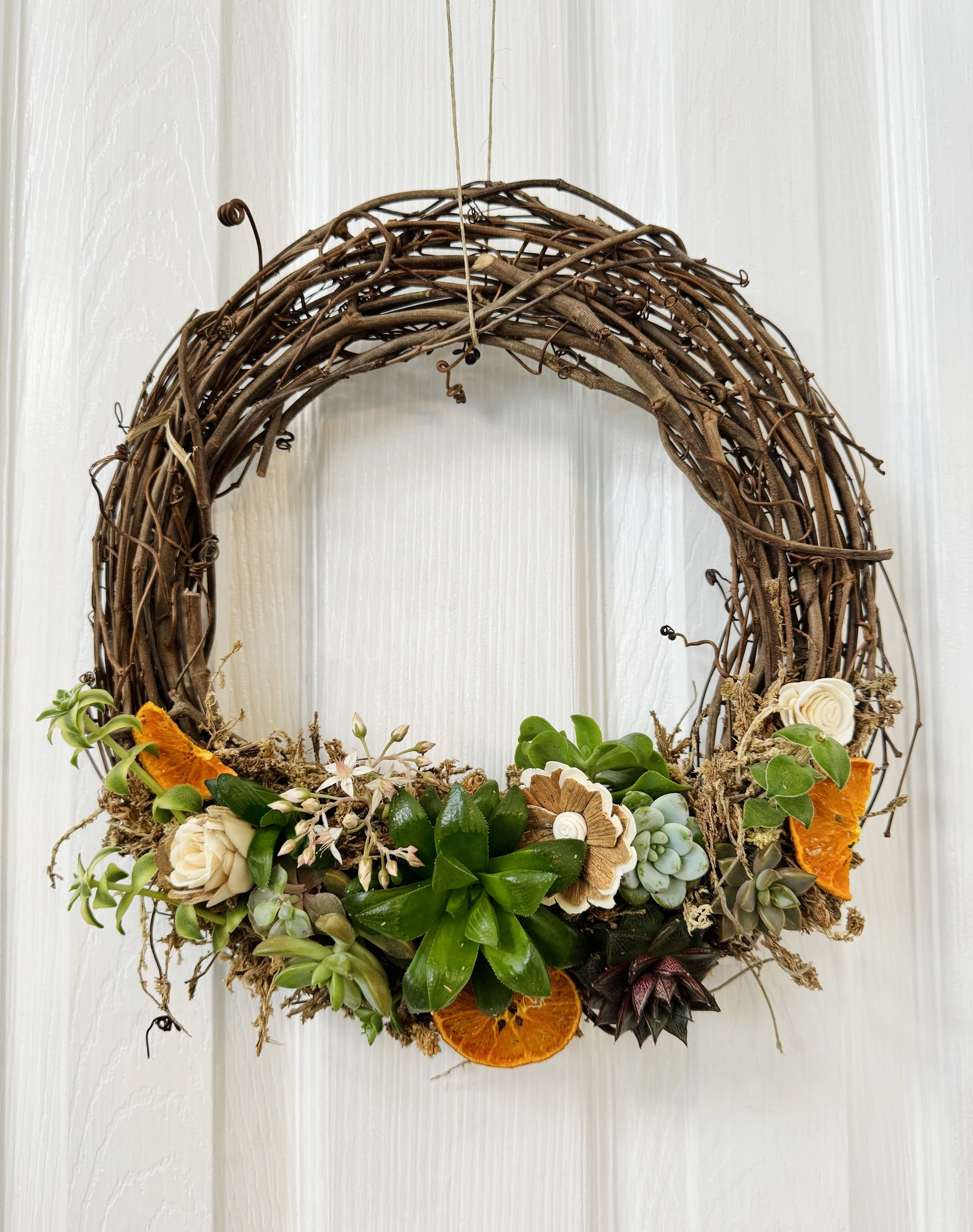 A decorative wreath made of intertwined brown twigs, decorated with succulents, dried orange slices, and flowers, hanging on a white wooden door.