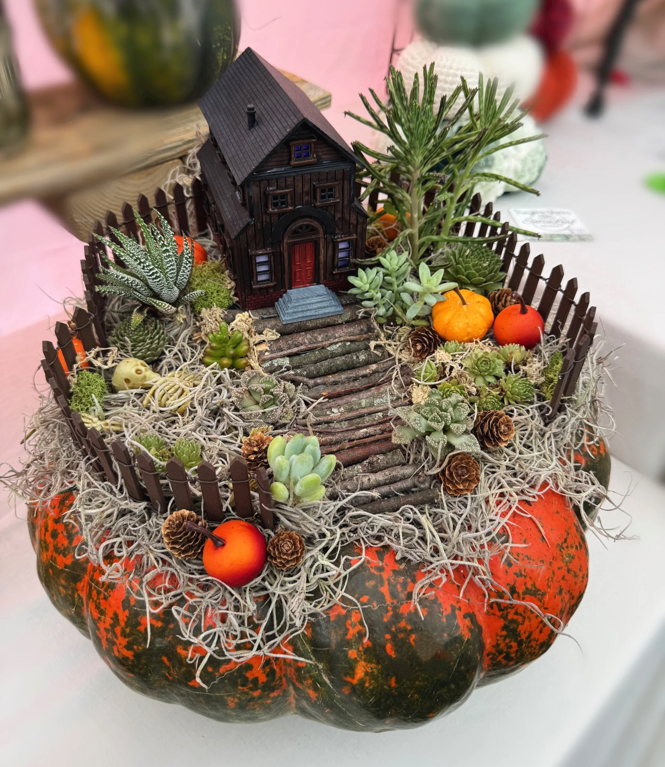 Decorative miniature scene with a small house, succulents, orange pumpkins, pinecones, and twigs on a speckled pumpkin.