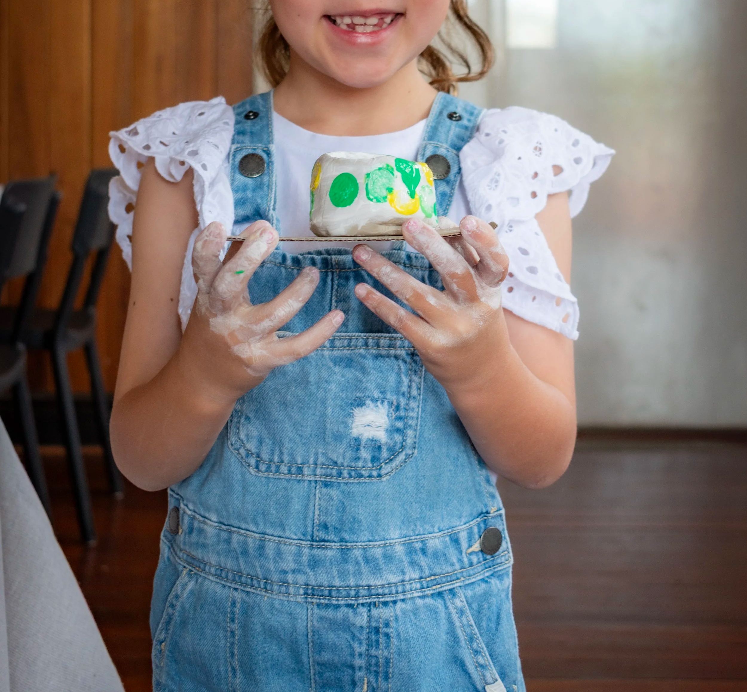 Young girl holding a painted rock with colorful circles, wearing a white eyelet top and denim overalls, smiling.