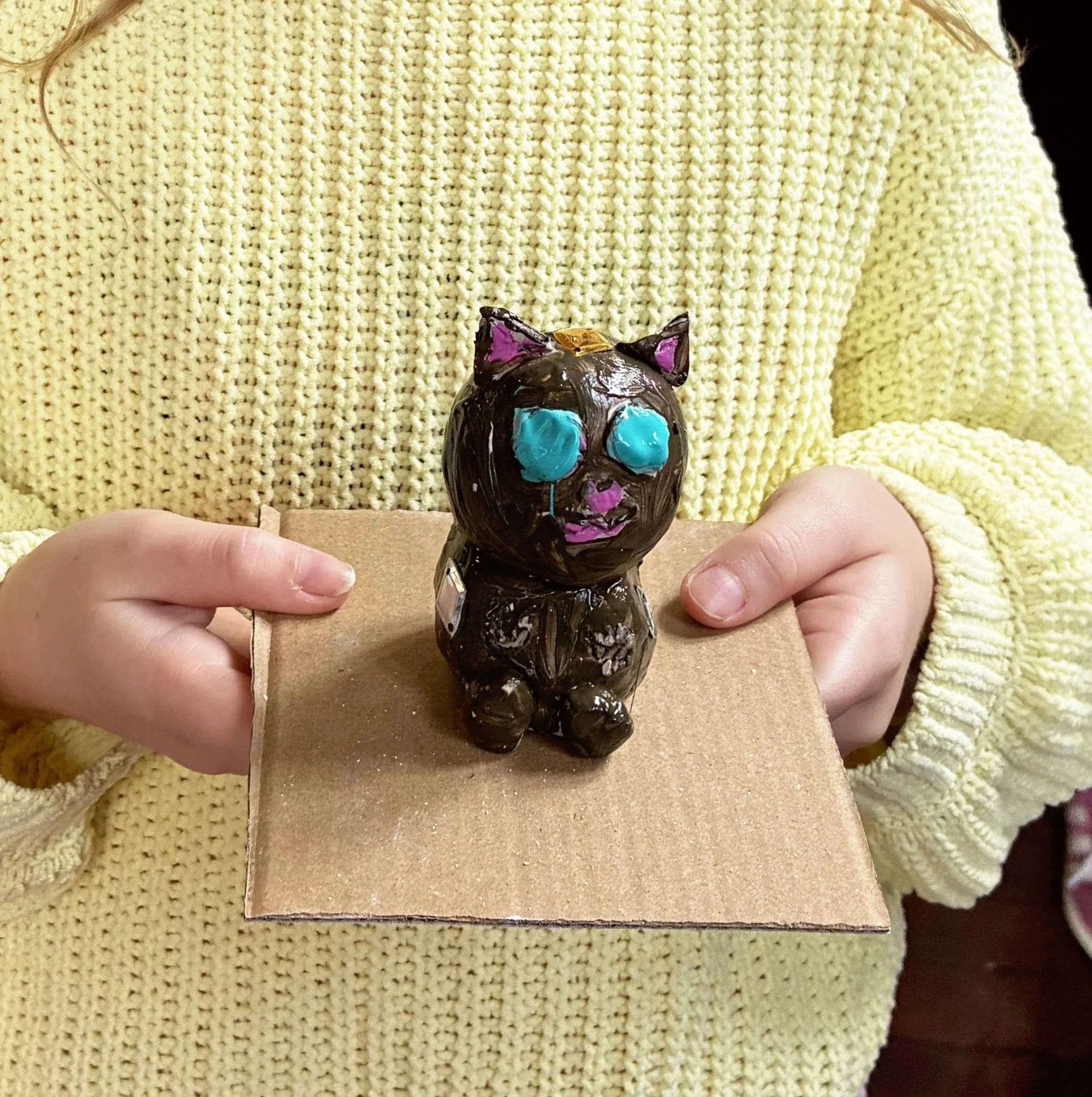A person holding a small, hand-painted figurine of a black cat with purple ears, blue eyes, and pink accents on a beige surface, dressed in a yellow knitted sweater.