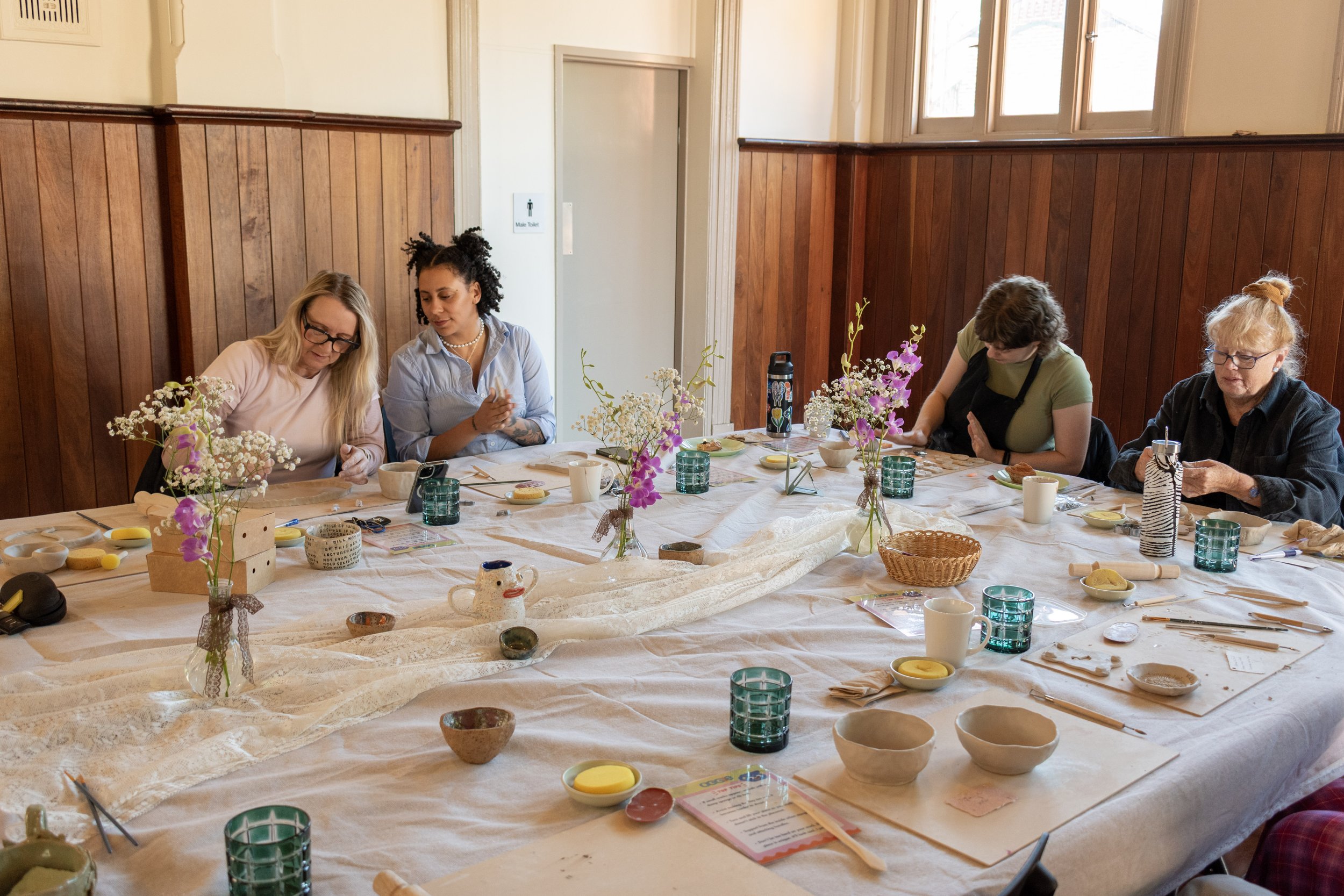 A group of people sitting around a table set with various bowls, cups, and flowers in vases, likely preparing for a meal or crafting activity in a room with wooden paneling and large windows.
