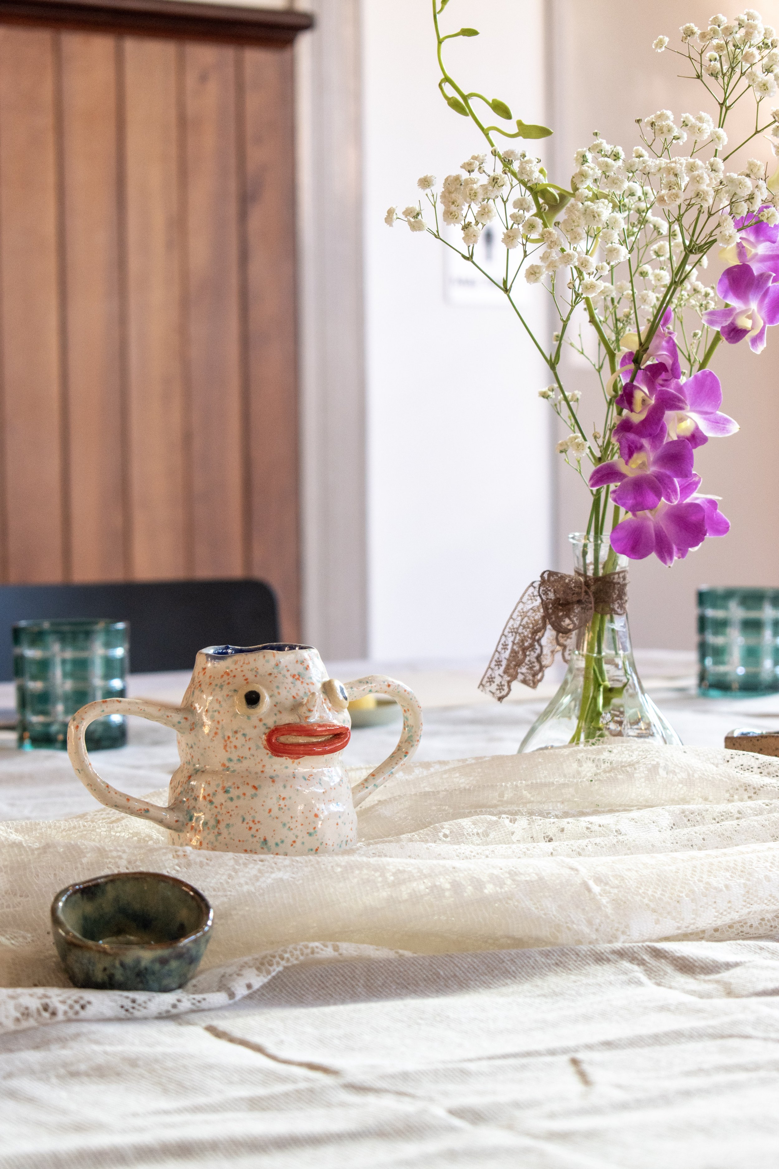 Vase with white and purple flowers on a table with various ceramic items, set in a room with wooden furniture.