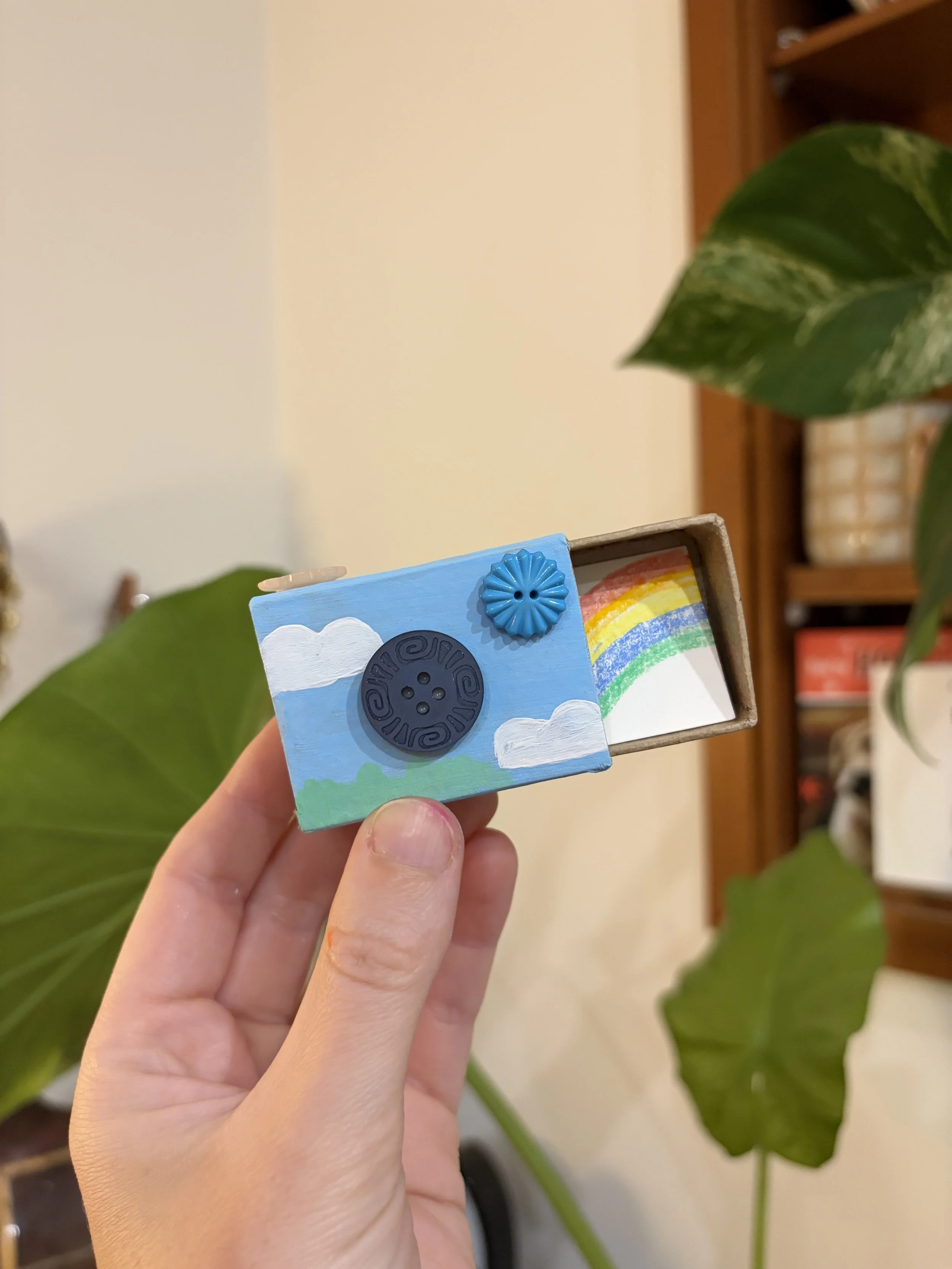 A small, hand-held painted wooden box featuring clouds, a rainbow, and buttons as design elements.