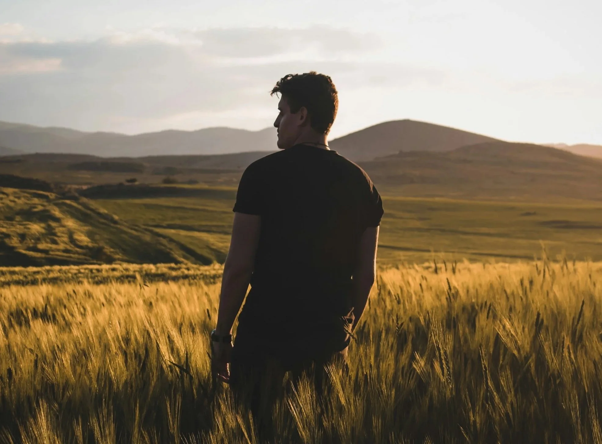 A man standing outdoors in quiet reflection, facing away toward the trees