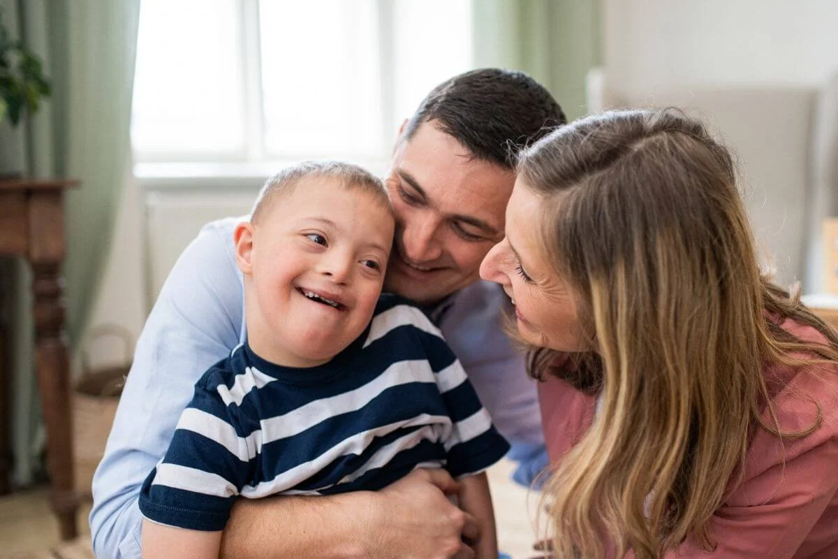 A joyful family of three, including a smiling young boy in a striped shirt, being embraced by a man and a woman in a cozy indoor setting, all displaying warm expressions of happiness.