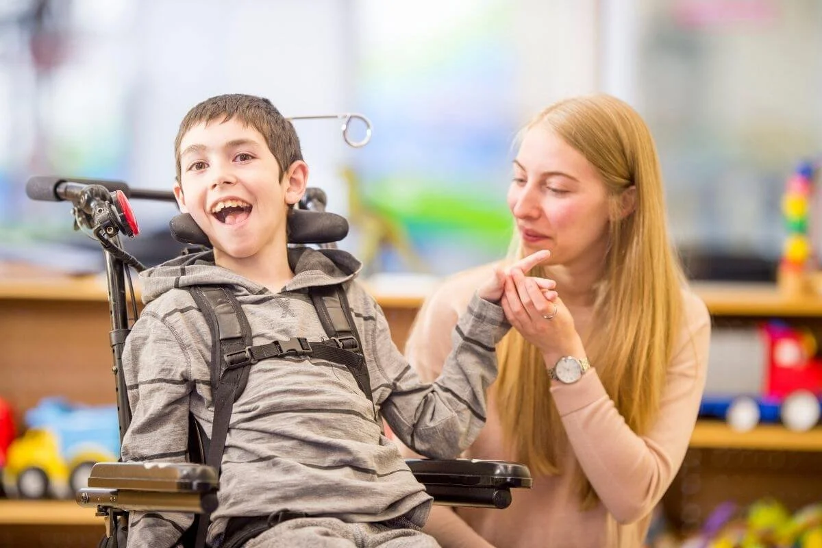 A young boy in a wheelchair smiling, holding hands with a woman with long red hair who is smiling and looking at him, in a room with colorful toys.