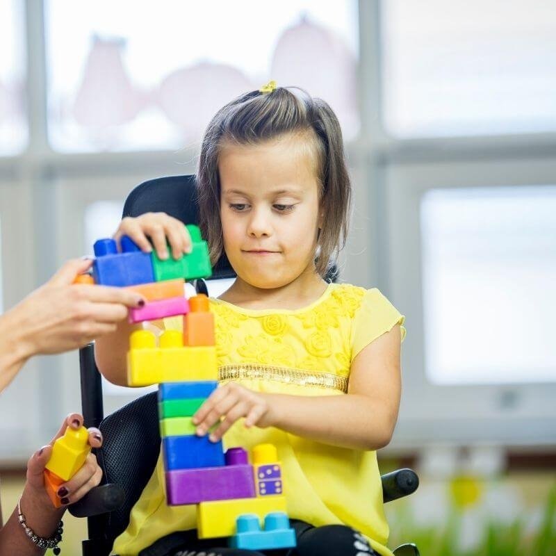 Young girl in yellow dress stacking colorful toy blocks indoors.
