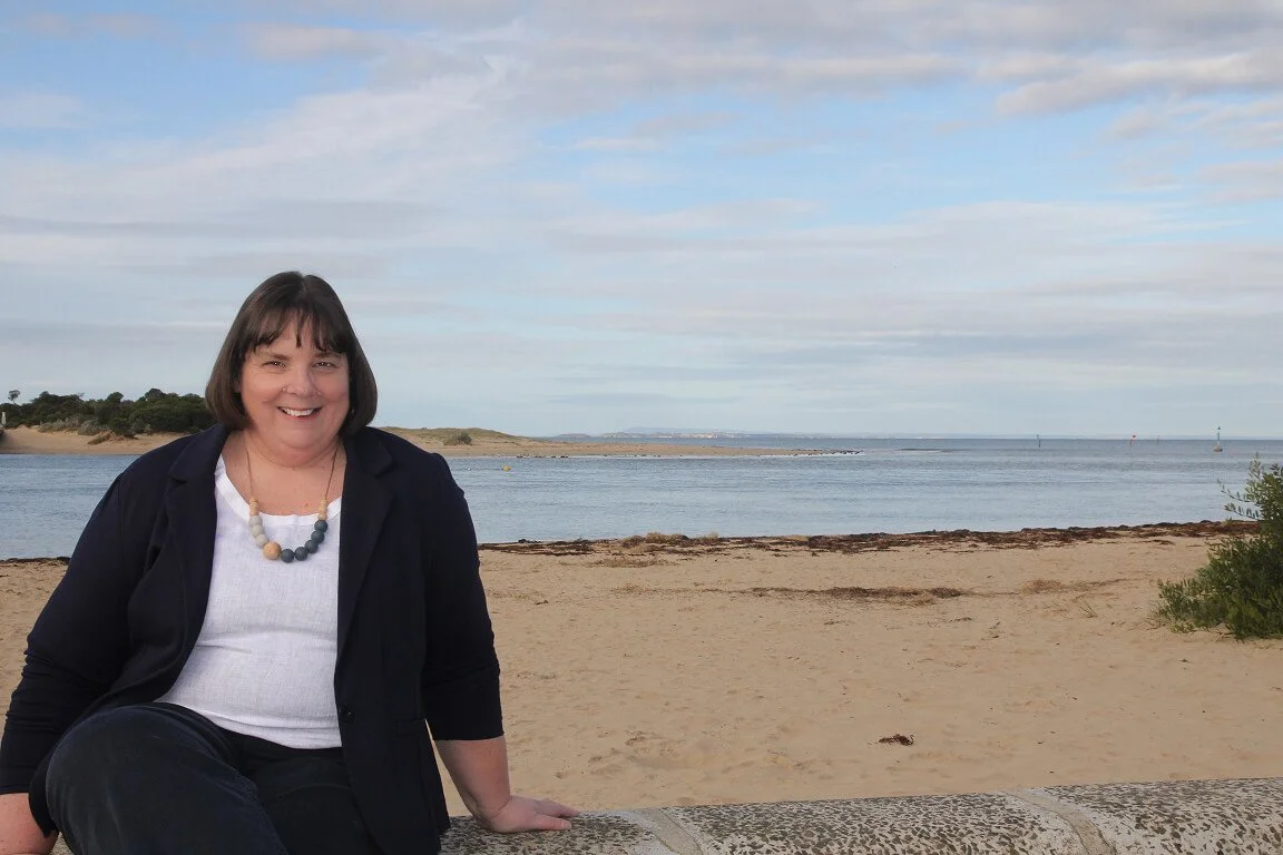 Smiling woman sitting on a stone ledge at the beach with sandy shoreline, water, and partly cloudy sky in the background.
