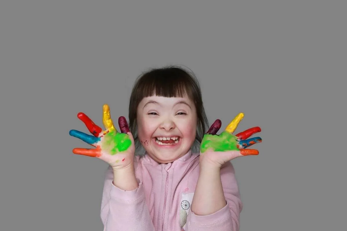 A young girl with a big smile shows her hands covered in colorful finger paint, with bright colors on her fingers and palms, holding her hands up in front of her face against a plain gray background.
