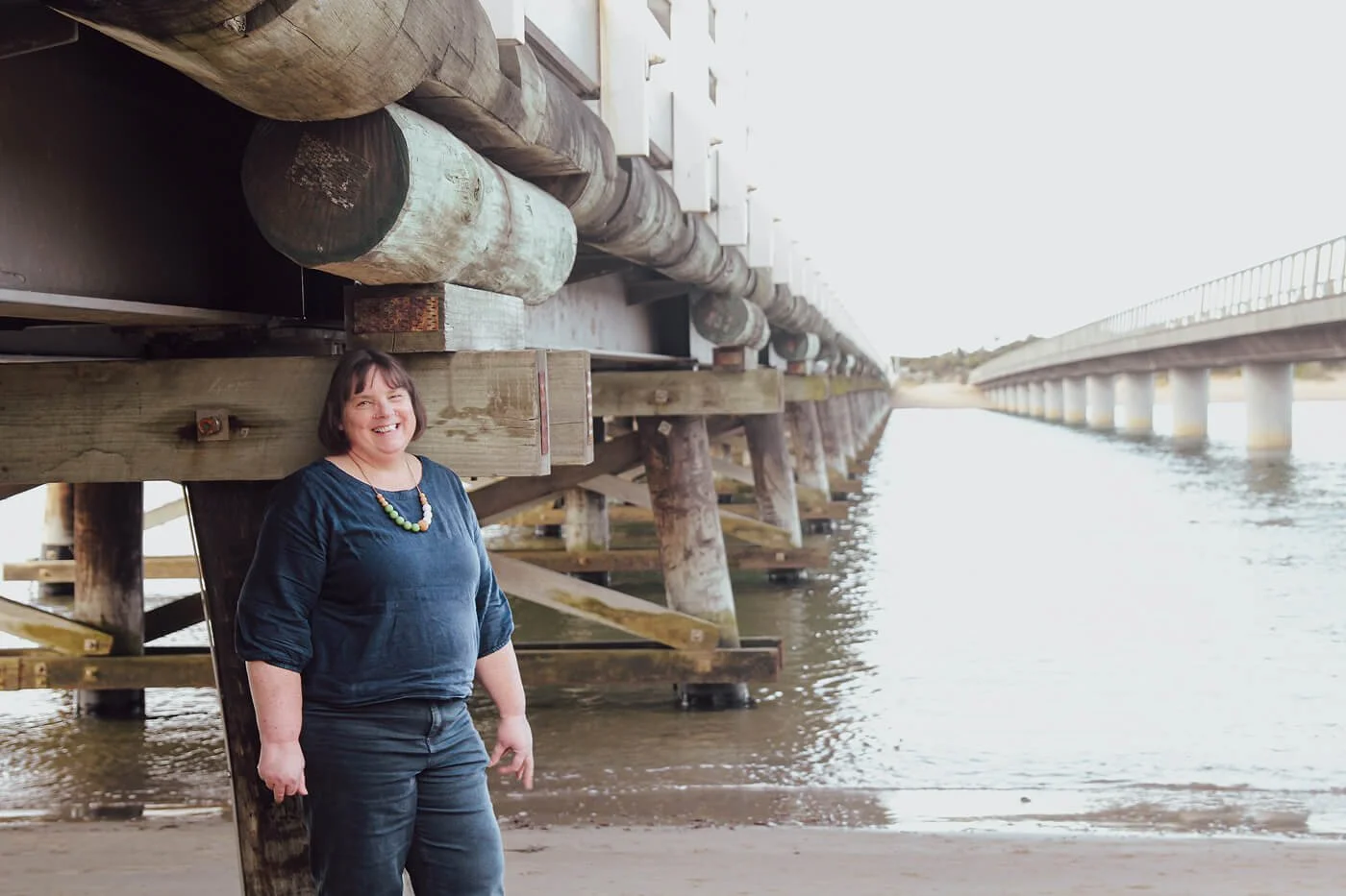 A woman standing under a wooden pier on a beach, smiling, with water and the continuation of the pier in the background.