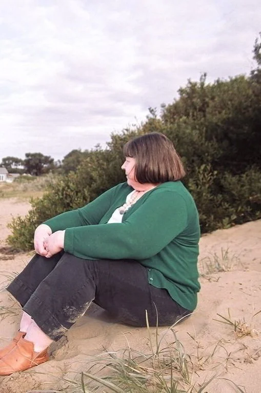 A woman with short brown hair sits on sandy ground surrounded by greenery, looking to the left and smiling.