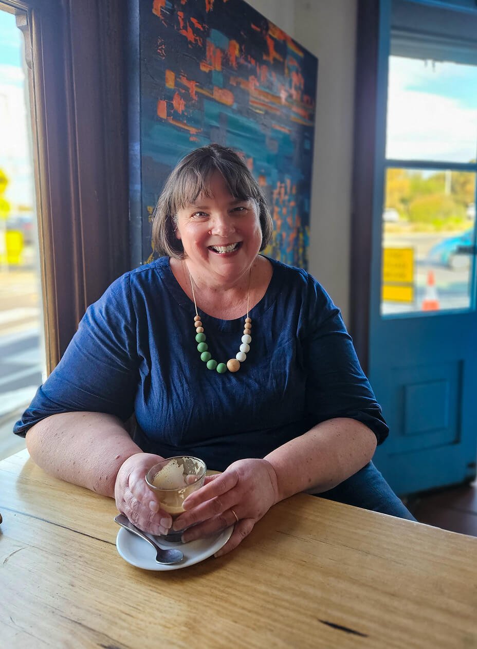 A woman with short brown hair smiling and sitting at a wooden table with a cup in her hands, inside a cafe with colorful artwork and large windows showing a street view.