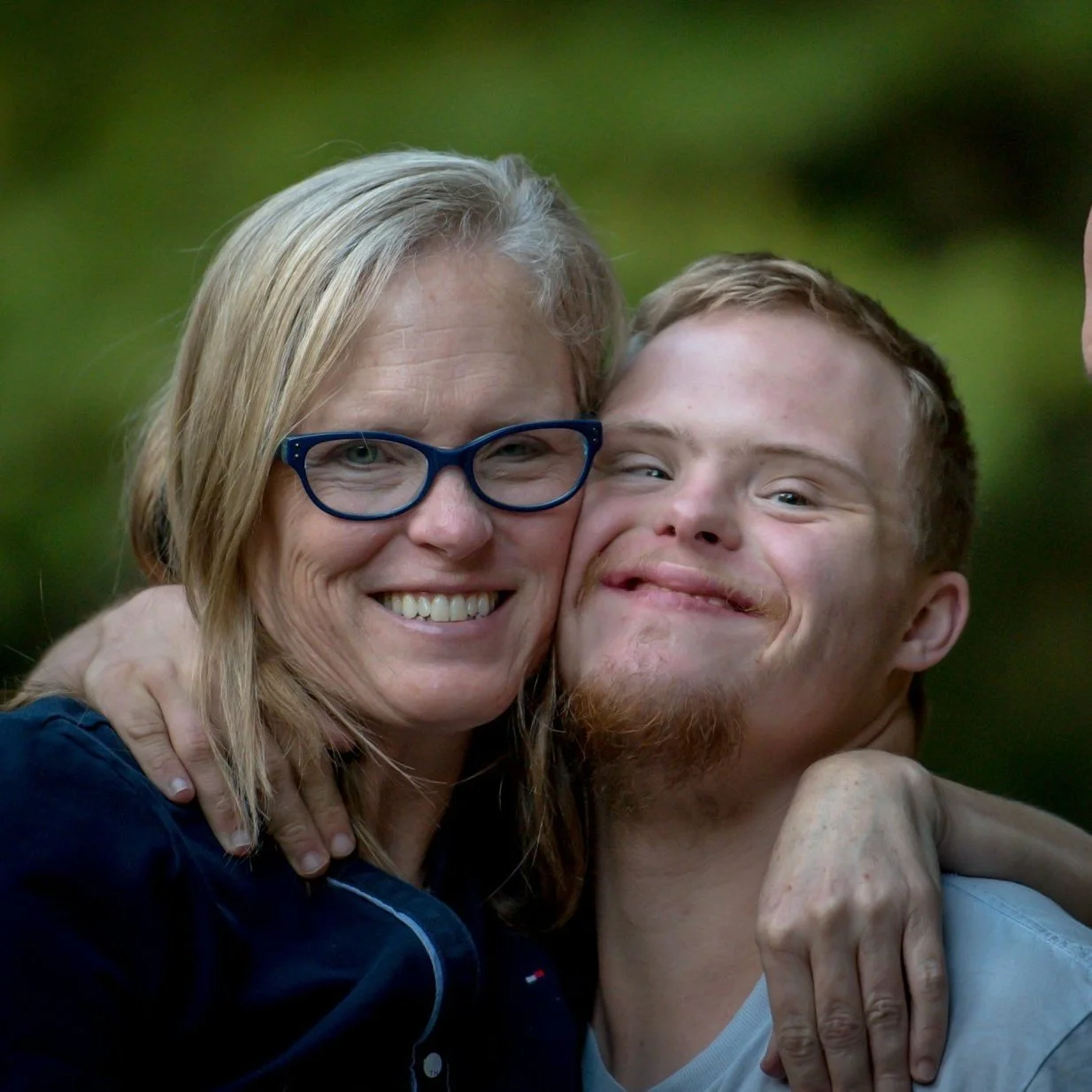 A woman with blonde hair, glasses, and a navy jacket hugging a young man with short hair, a beard, and a light blue shirt, both smiling and looking happy.