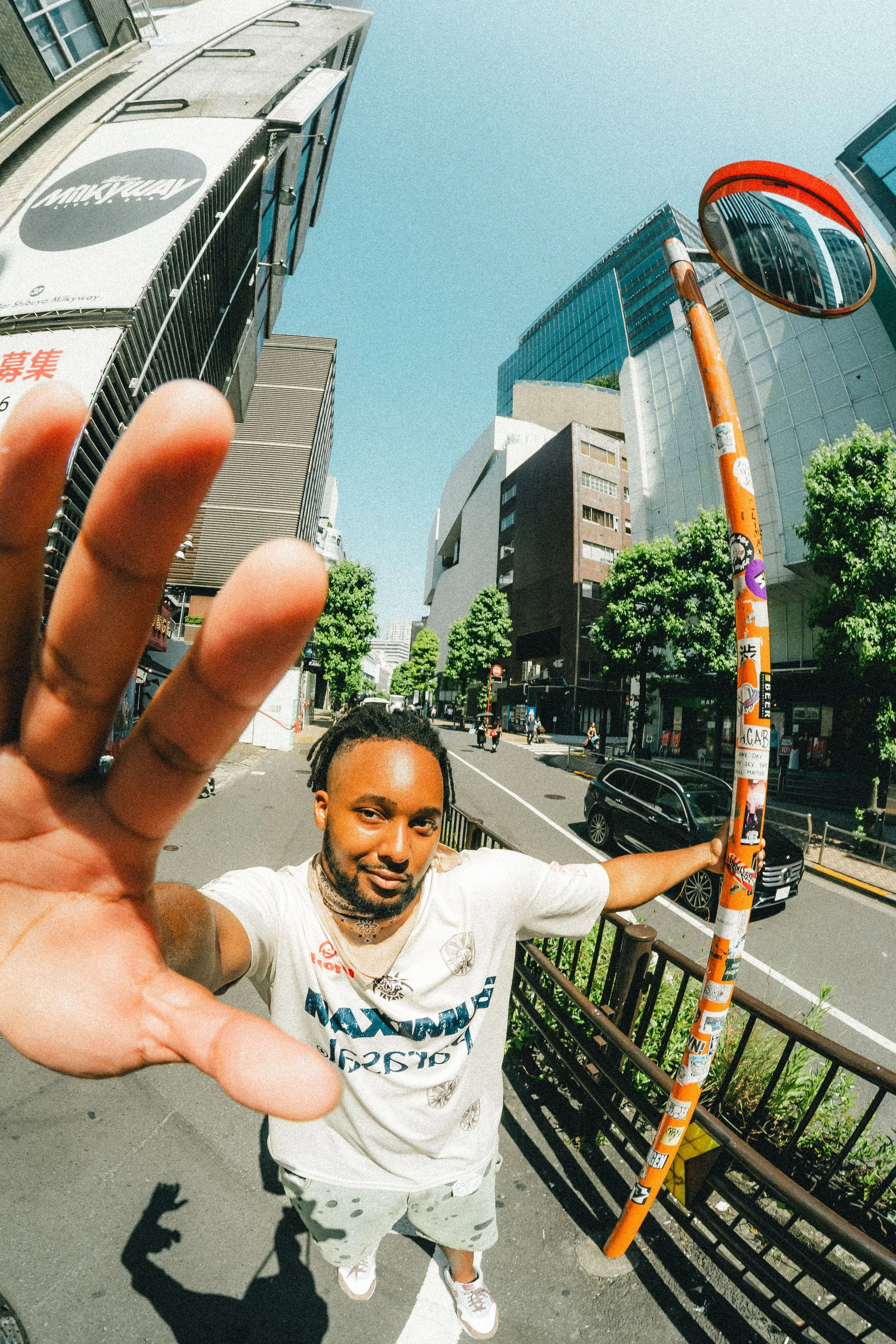 A man reaching towards the camera with a street scene behind him, including buildings, trees, cars, a street mirror, and a sunny sky.