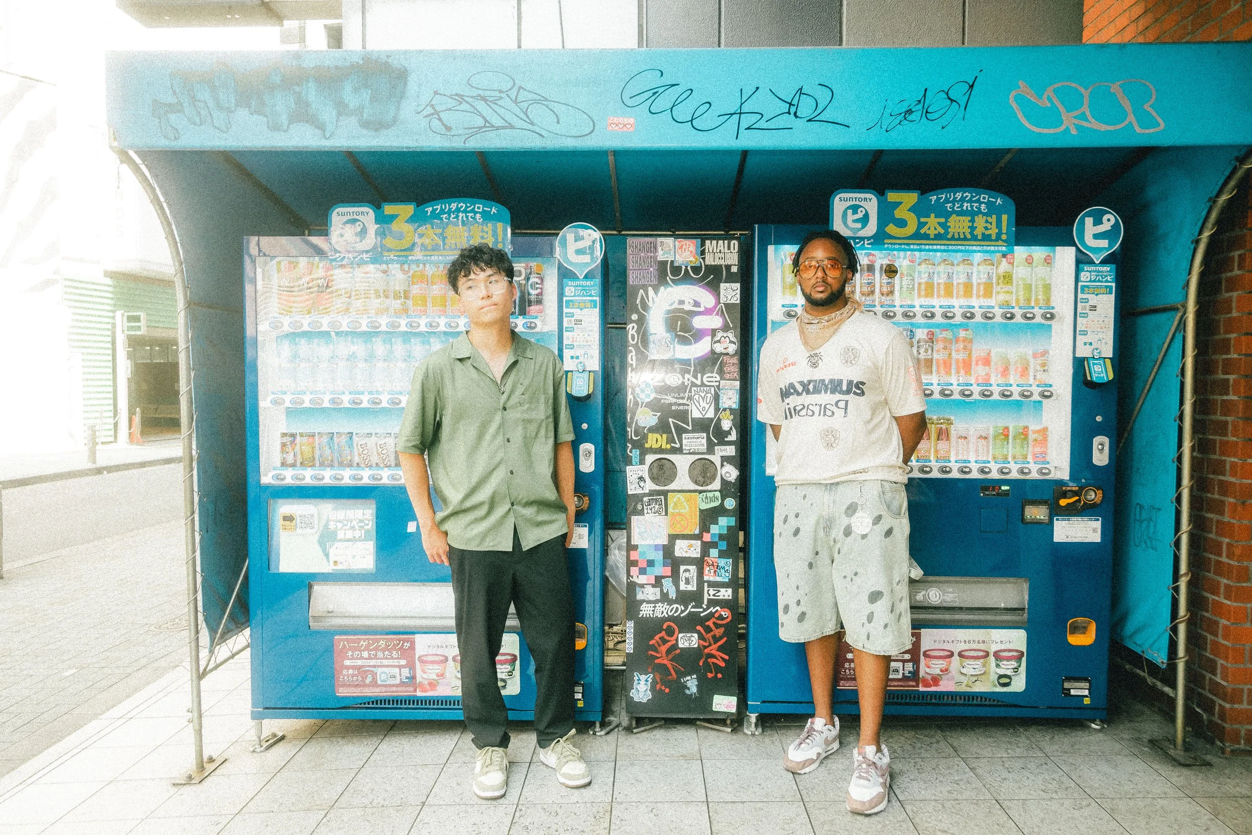 Two men standing in front of vending machines outside. One is wearing a green shirt and black pants, the other is dressed in a light t-shirt and shorts with glasses.
