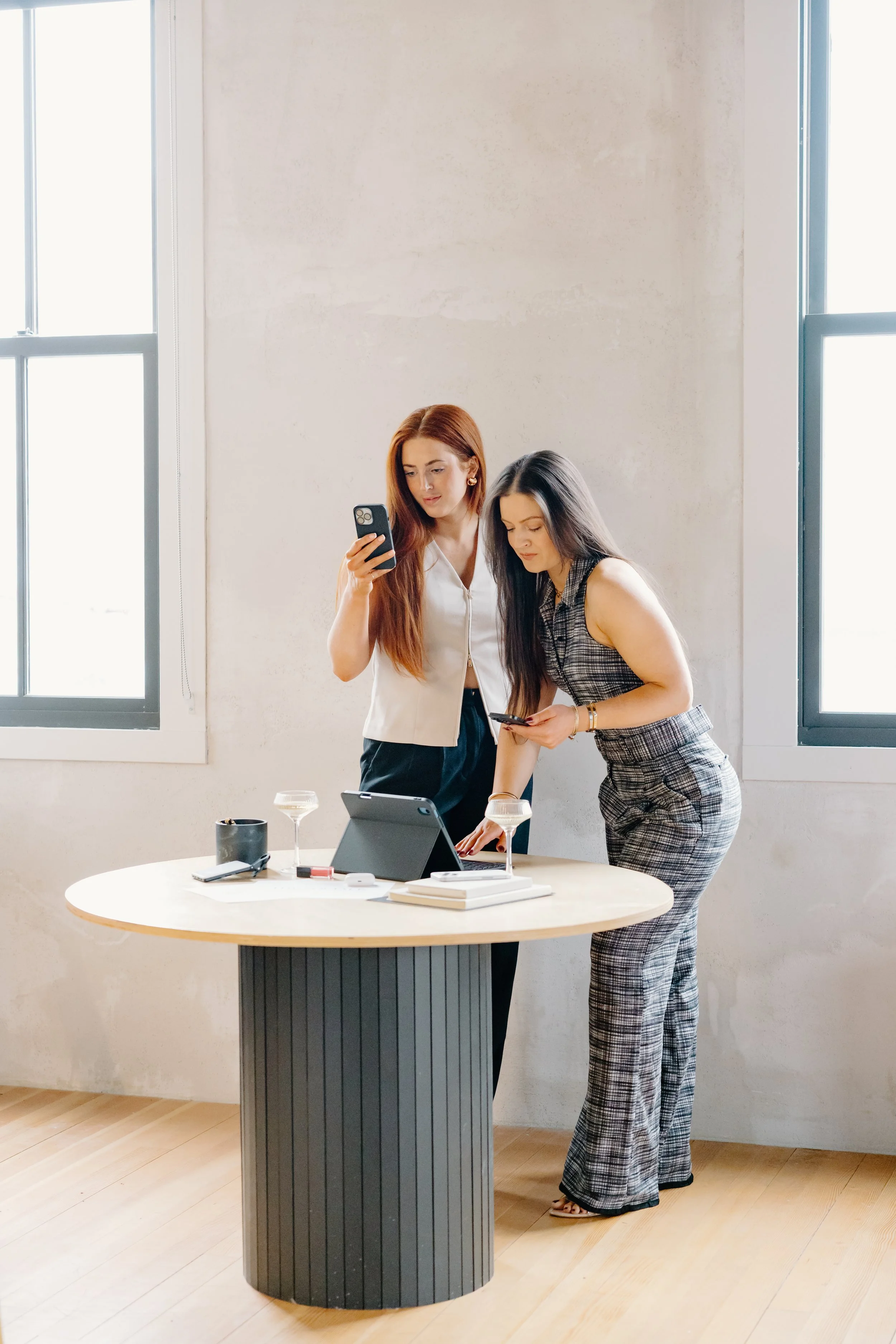 Two women in business attire standing at a round table with a tablet, two glasses of wine, and office supplies, in a bright room with large windows.