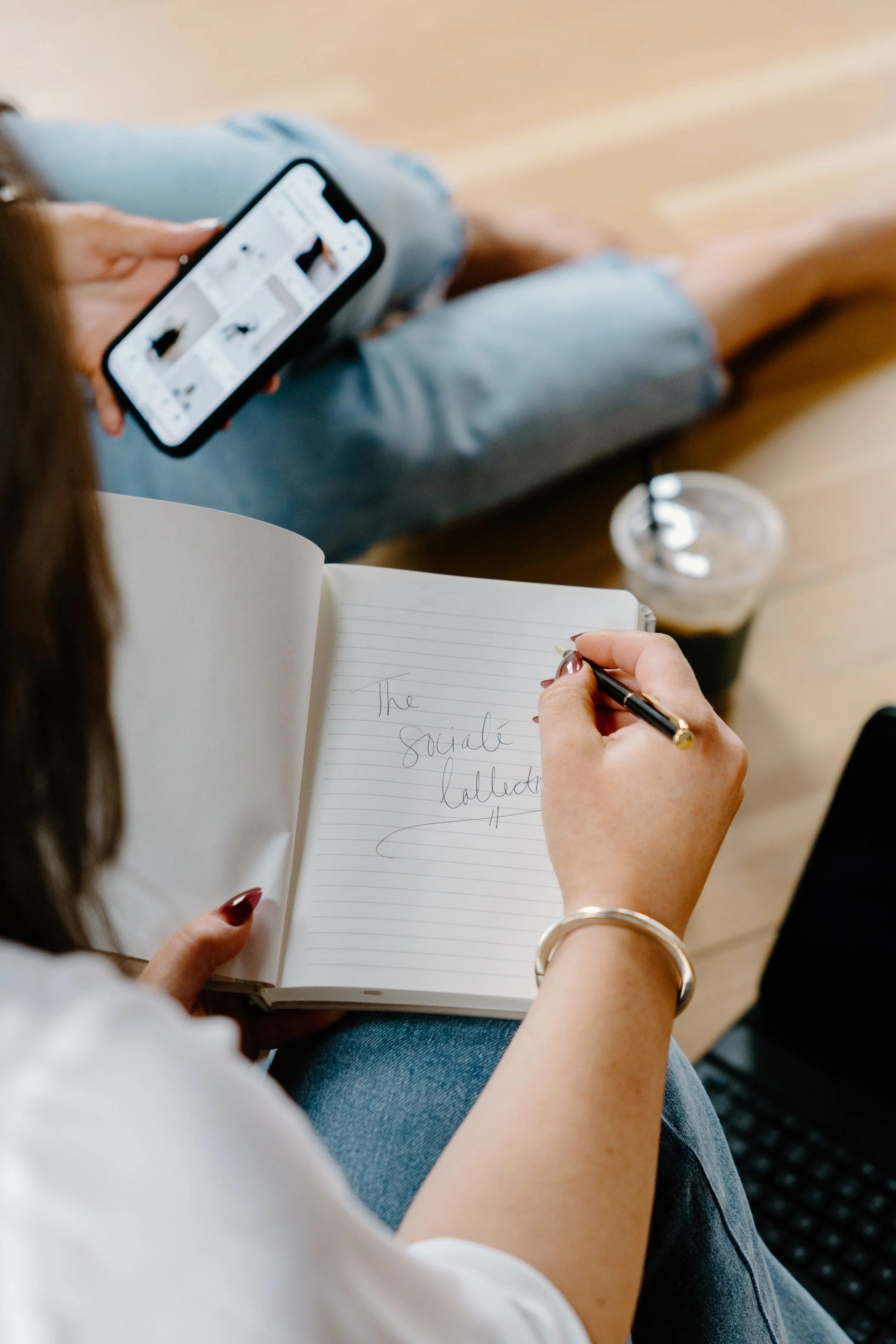 A person sitting down, holding a phone in one hand and an open notebook in the other, writing with a pen. The notebook has the words 'The sociale collection' written on the page. There is a plastic cup with a dark beverage and a straw on the table nearby.