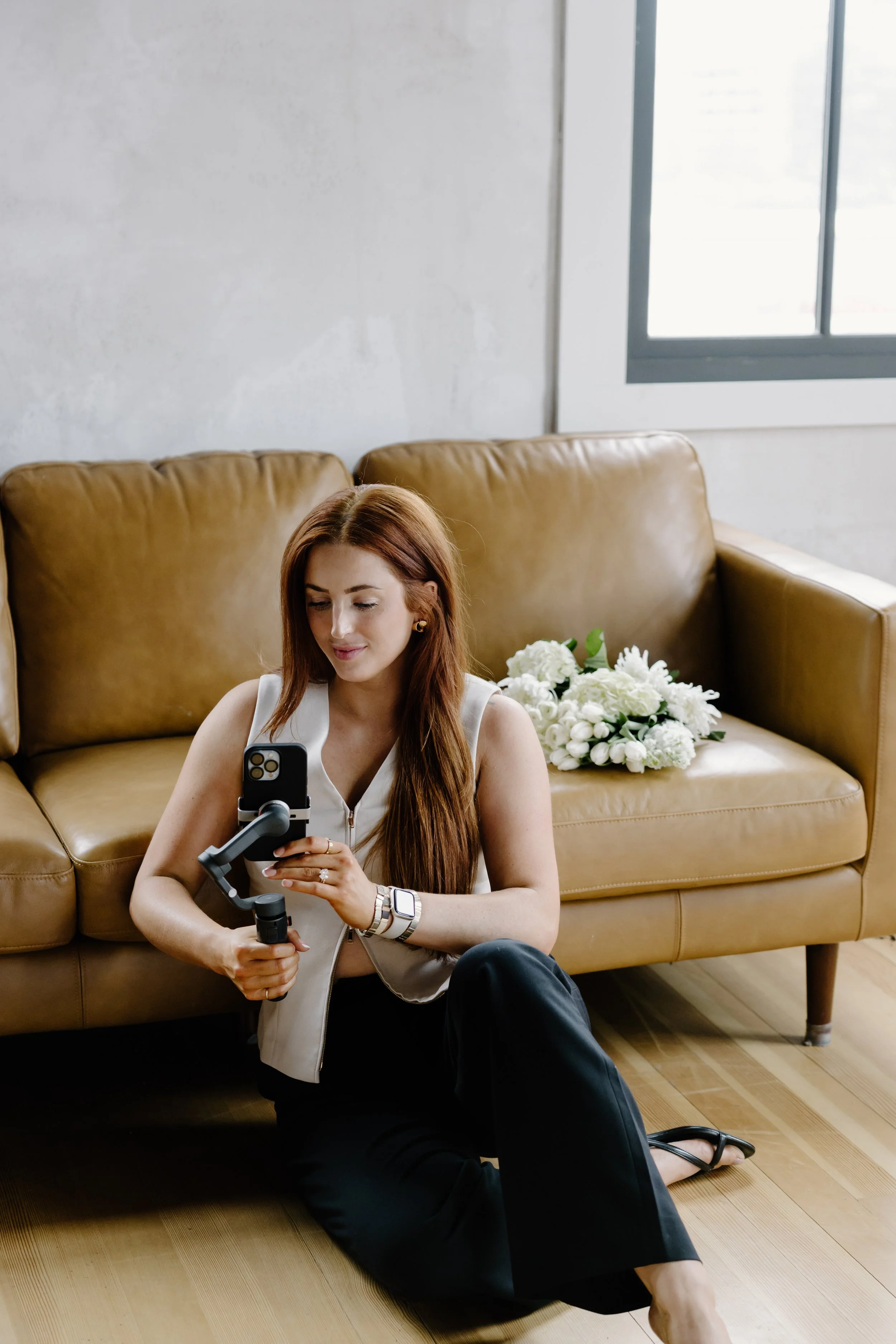 A woman with long red hair sitting on a hardwood floor in front of a tan leather couch, holding a camera stabilizer and smiling at her phone. A bouquet of white flowers is on the couch, and a window with gray framing is in the background.